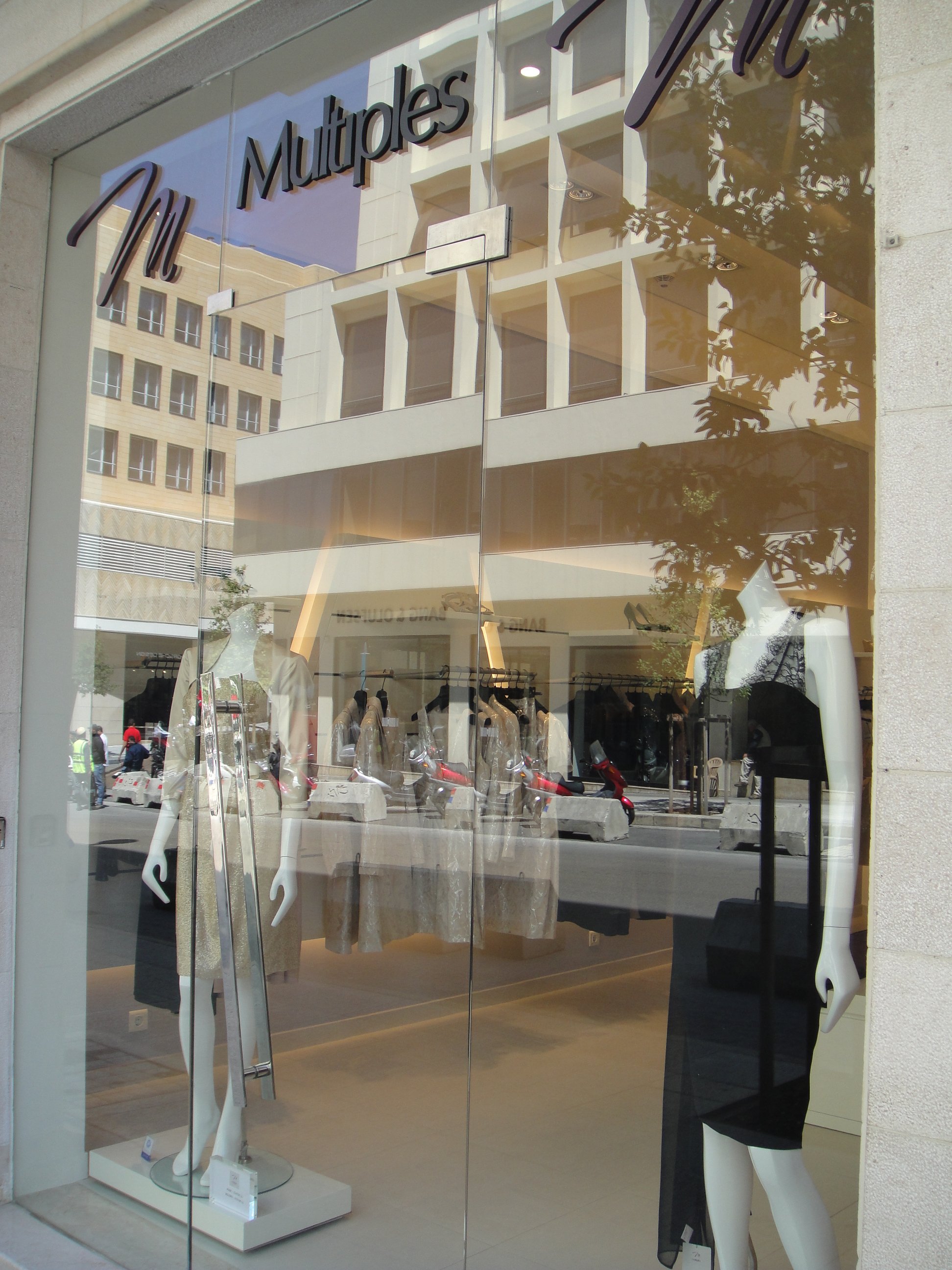 Store window display with mannequins wearing clothing, reflected building and trees outside.