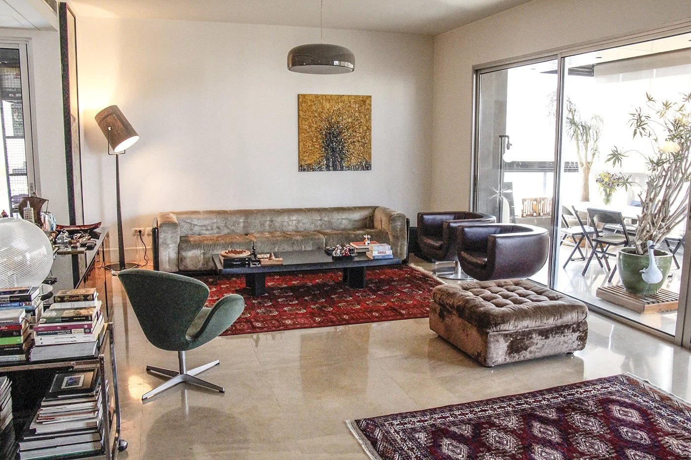 Living room with a beige velvet sofa, two dark brown armchairs, and a brown tufted ottoman. A black coffee table has books and decorative items. A red patterned rug covers the floor. A modern floor lamp and an art piece are on the wall. Sliding glass