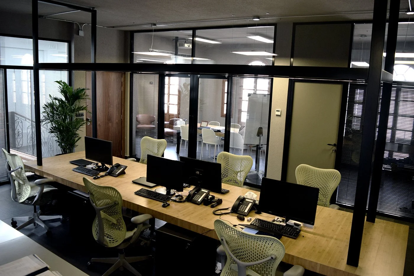 Office conference room with glass walls, a wooden table with computers, phones, and office chairs, and a meeting room in the background with additional chairs and tables.