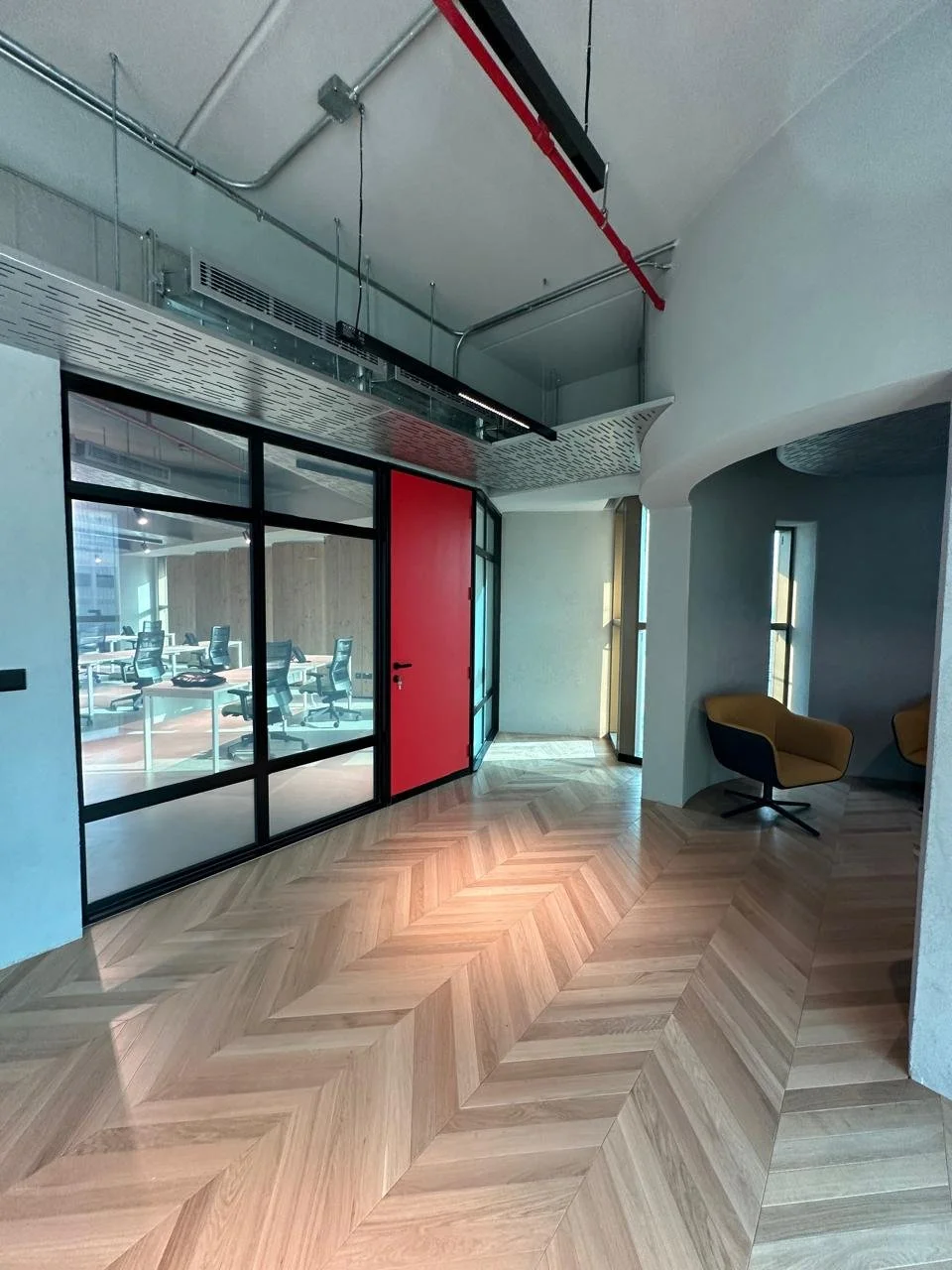 Modern office interior with wooden herringbone patterned flooring, a glass-walled conference room, a red door, and a seating area with yellow and black chairs near tall windows.