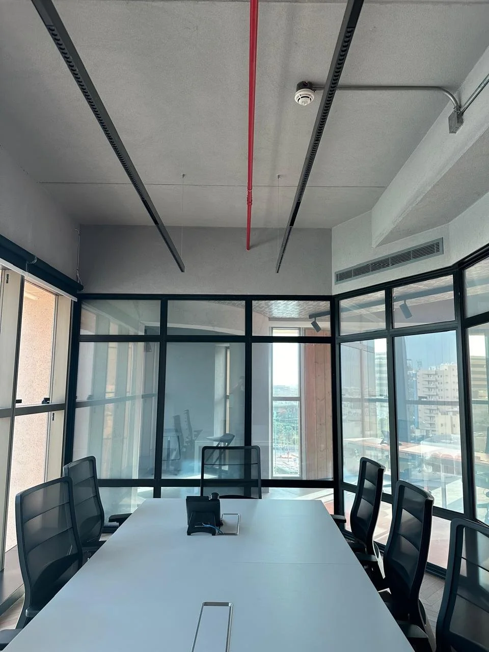 Empty conference room with a long white table, six black office chairs, glass walls, and city view outside.
