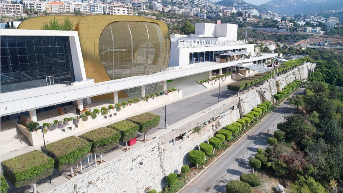 Aerial view of a modern casino building with a golden rounded structure and glass facade, situated on a hill with a landscaped walkway, trees, and a cityscape in the background.