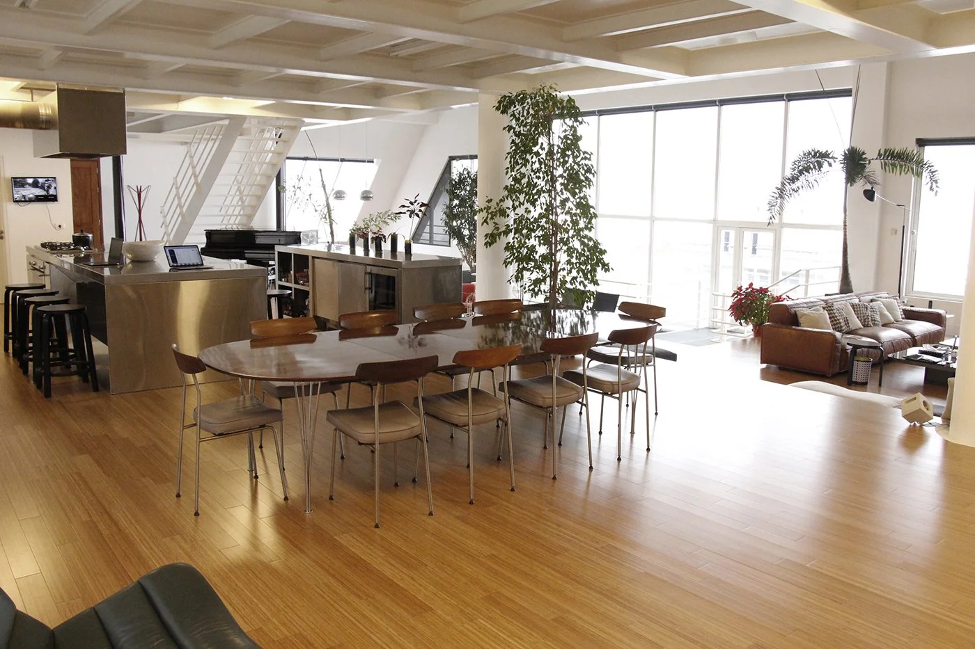 Open-plan living space with a large oval dining table with wooden chairs, a kitchen island with stools, a living area with a leather sofa, large windows with sunlight, and indoor plants.