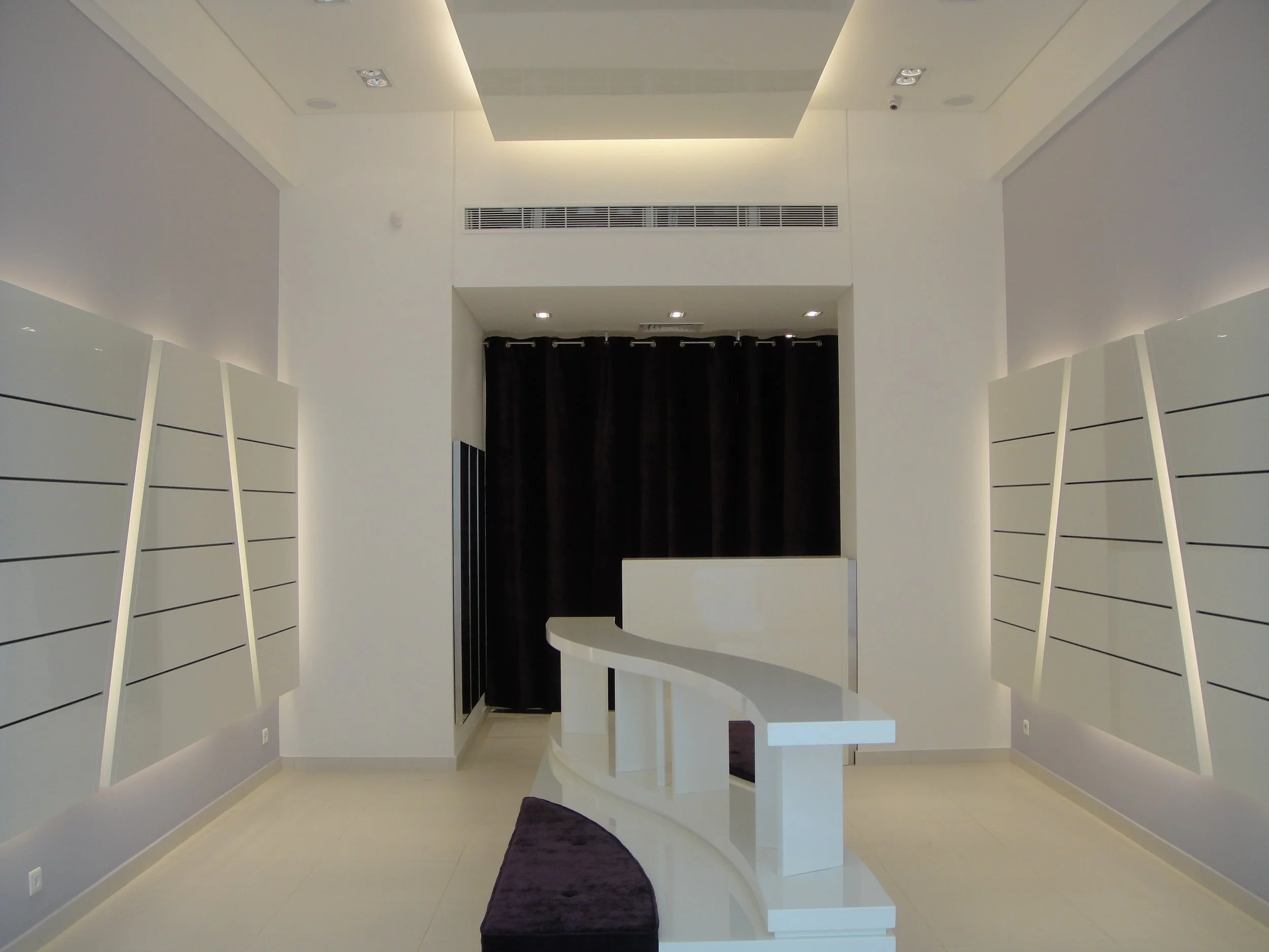 A modern, minimalist lobby or reception area with white walls, a white curved reception desk, a black curtain in the background, and sleek lighting fixtures on the ceiling.
