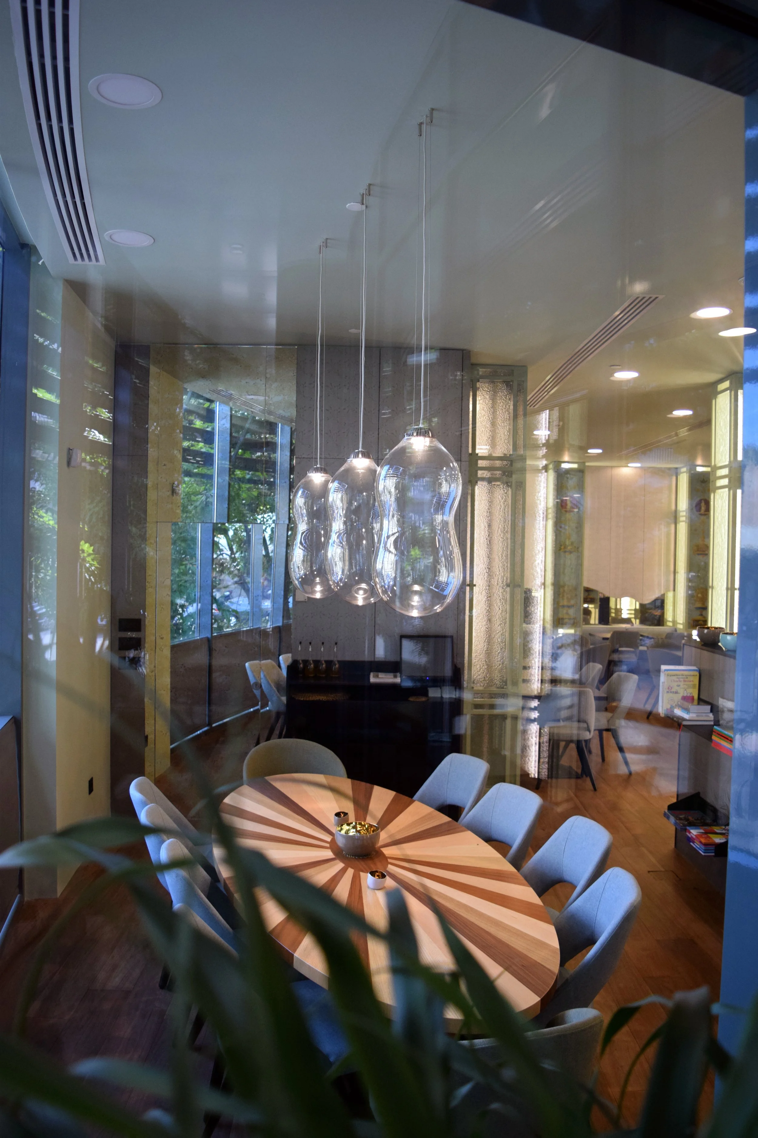 Interior view of a modern dining area with a wooden round table, white chairs, pendant lights, large windows, and a partial view of a cozy lounge.