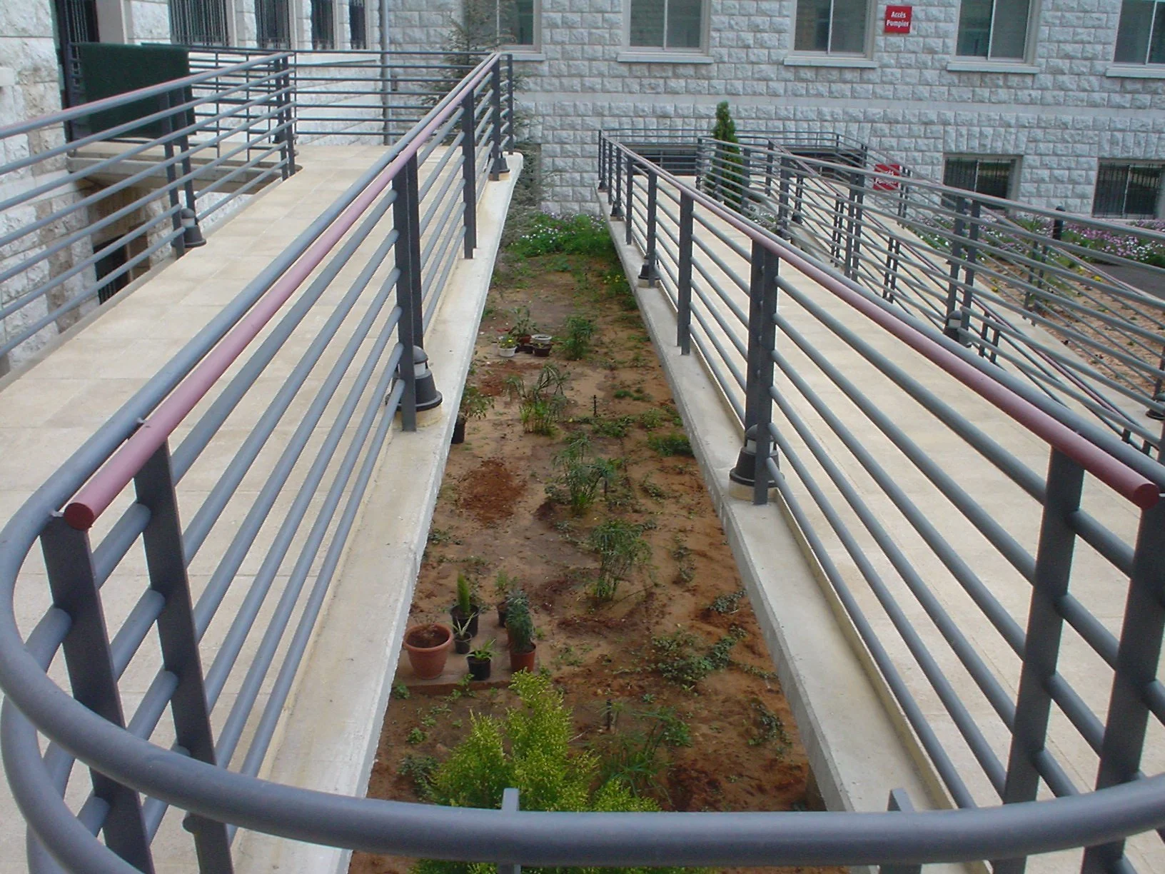 Apartment building exterior with a narrow garden bed between two walkways, potted plants and small shrubs planted in the soil.