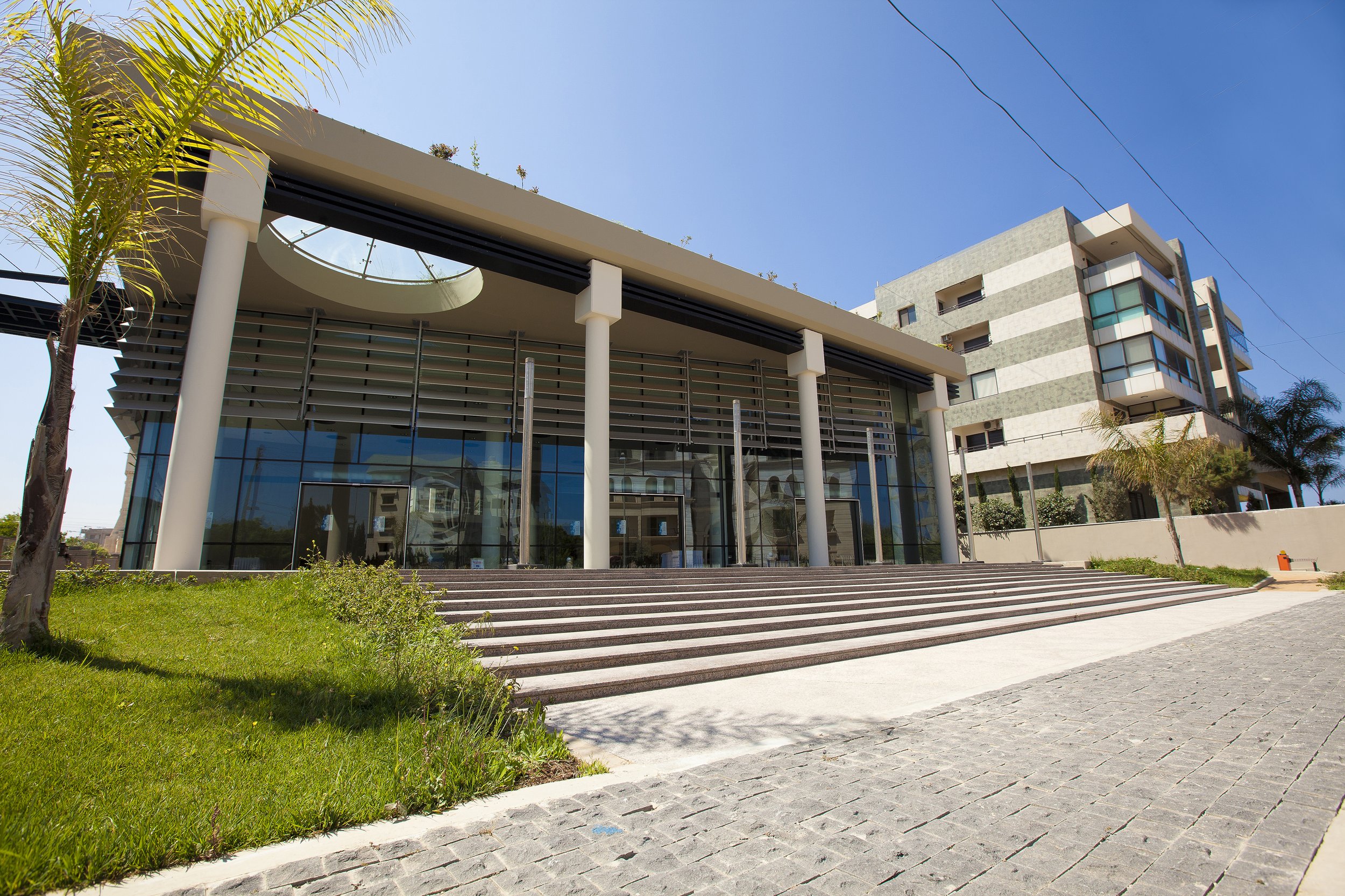 Modern building with glass facade and striped exterior, set against a clear blue sky, with a green lawn, trees, and wide steps at the entrance.