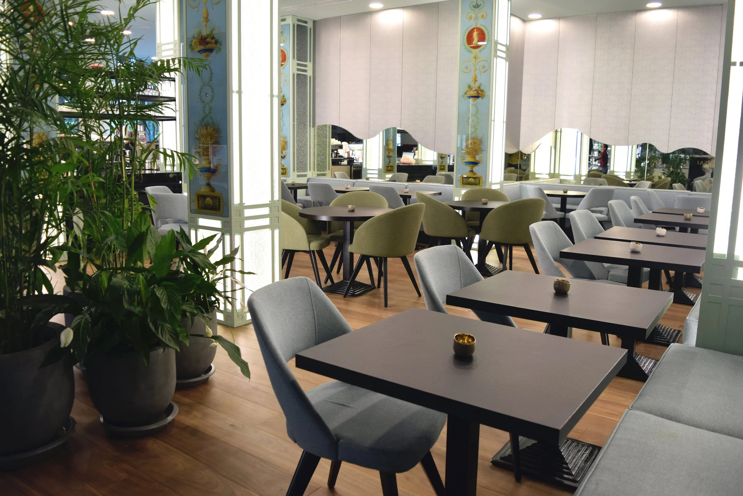 Empty restaurant with gray and green chairs around black tables, large potted plants on the left, decorative mirrors and paintings on the back wall, and a woman in a red shirt in the background.