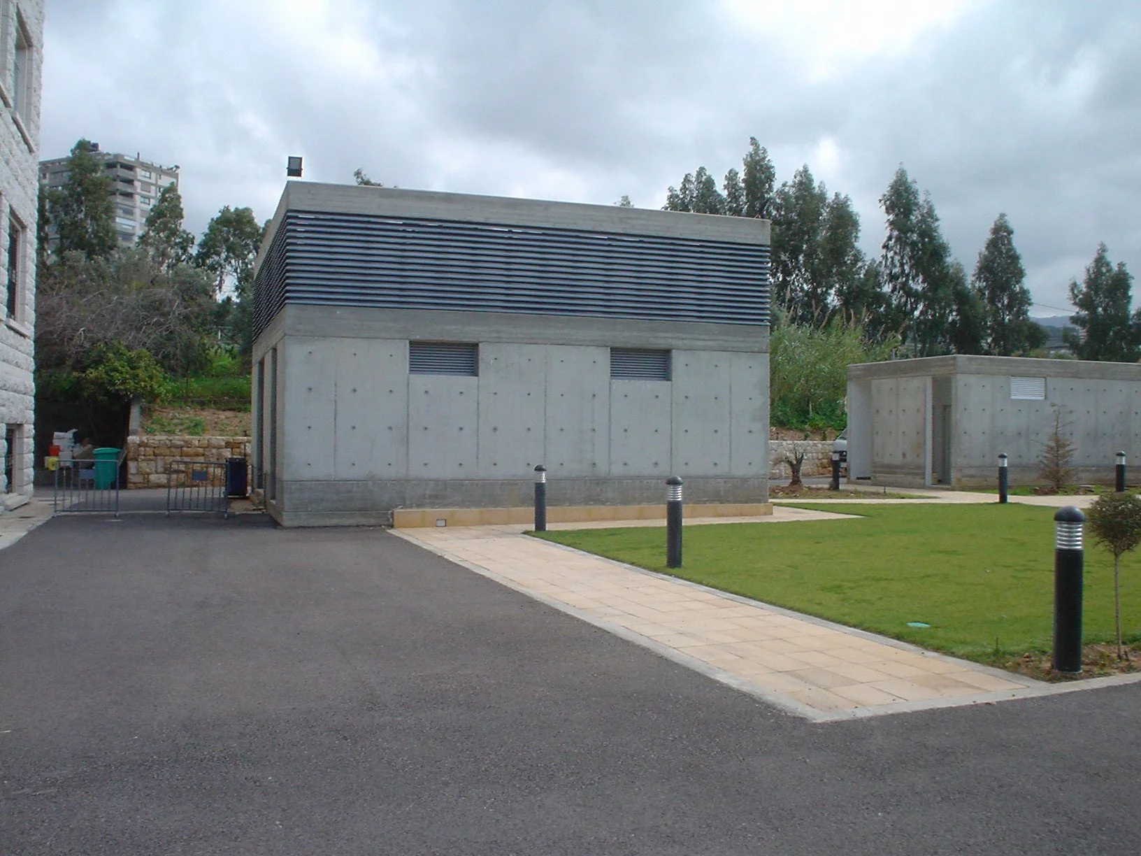 Two small concrete utility or storage buildings, one in the foreground and one in the background, with asphalt driveway, grass, and trees around, under cloudy sky.