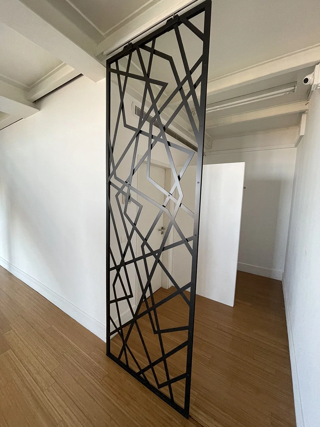 Decorative black metal partition with geometric pattern in a room with white walls and wooden flooring.