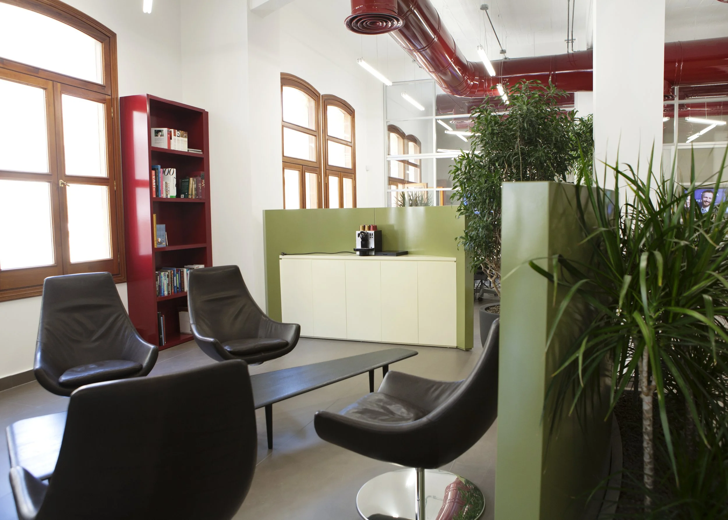 Modern office lounge with black leather chairs around a low coffee table, large windows with wooden frames, green plants, a white cabinet, and a coffee station on top, with exposed red ductwork and ceiling lights.