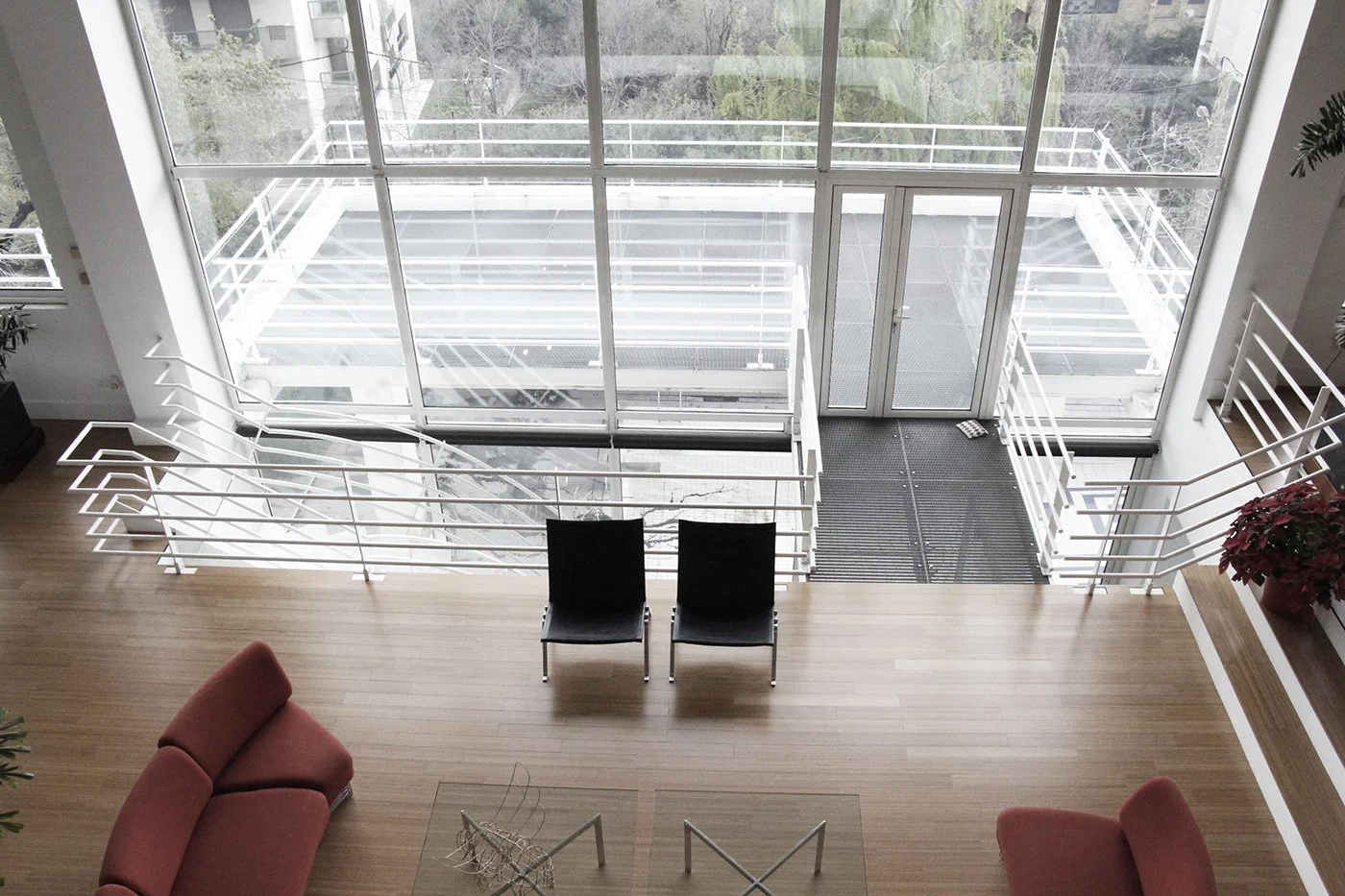 Interior of a modern apartment with a large glass window wall, two black chairs facing the window, a red sofa, and a wooden floor. Outside the window, there is a balcony with white railings and a view of trees.