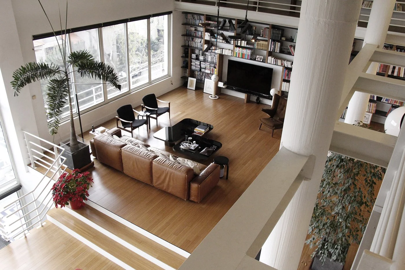 A spacious living room with a large window, wooden floors, a leather sofa, coffee table, black chairs, a TV on a wall with bookshelves, and indoor plants.