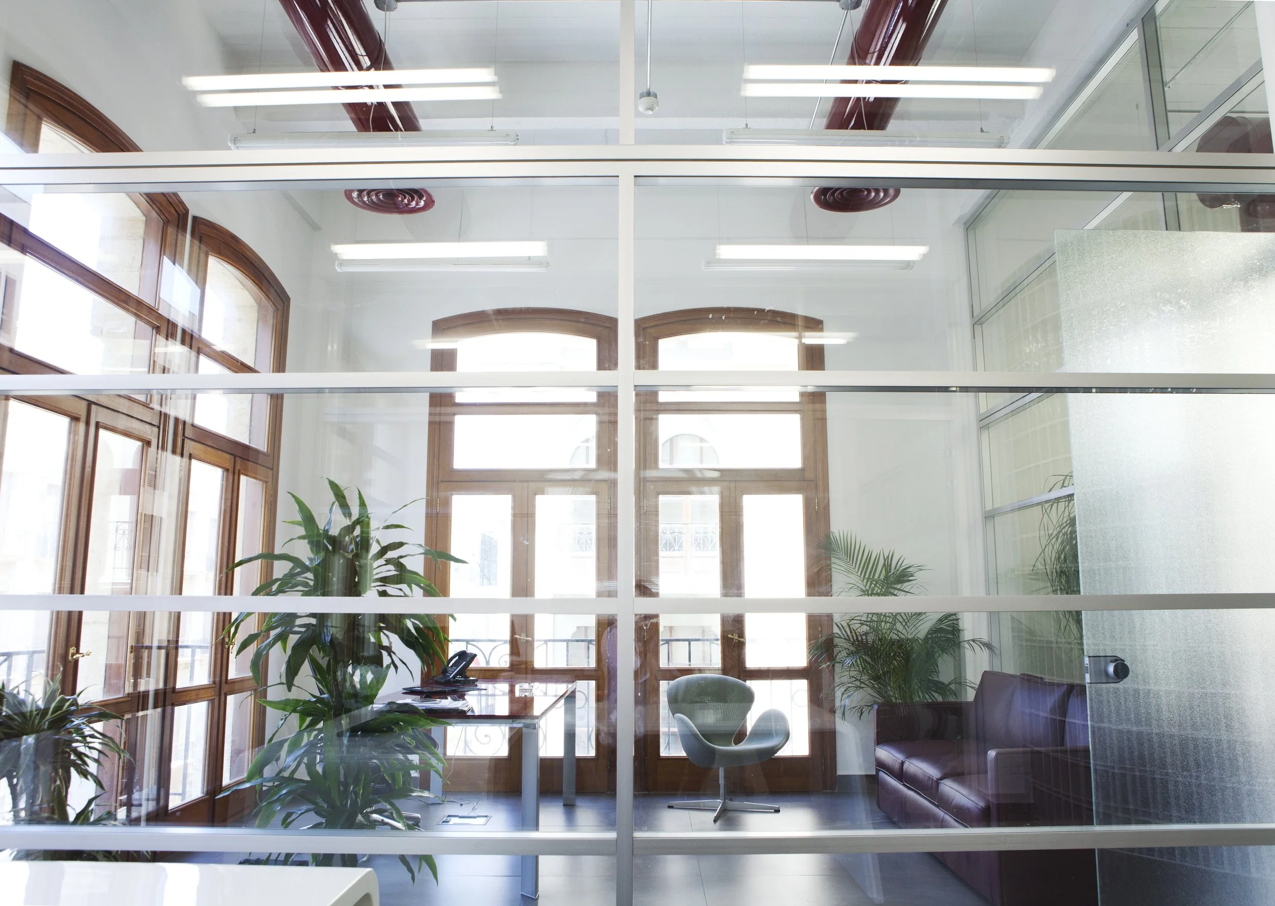 Office with glass walls, wooden doors, and large windows with natural light. There are green plants, a desk with a phone, a chair, a purple sofa, and a glass partition.
