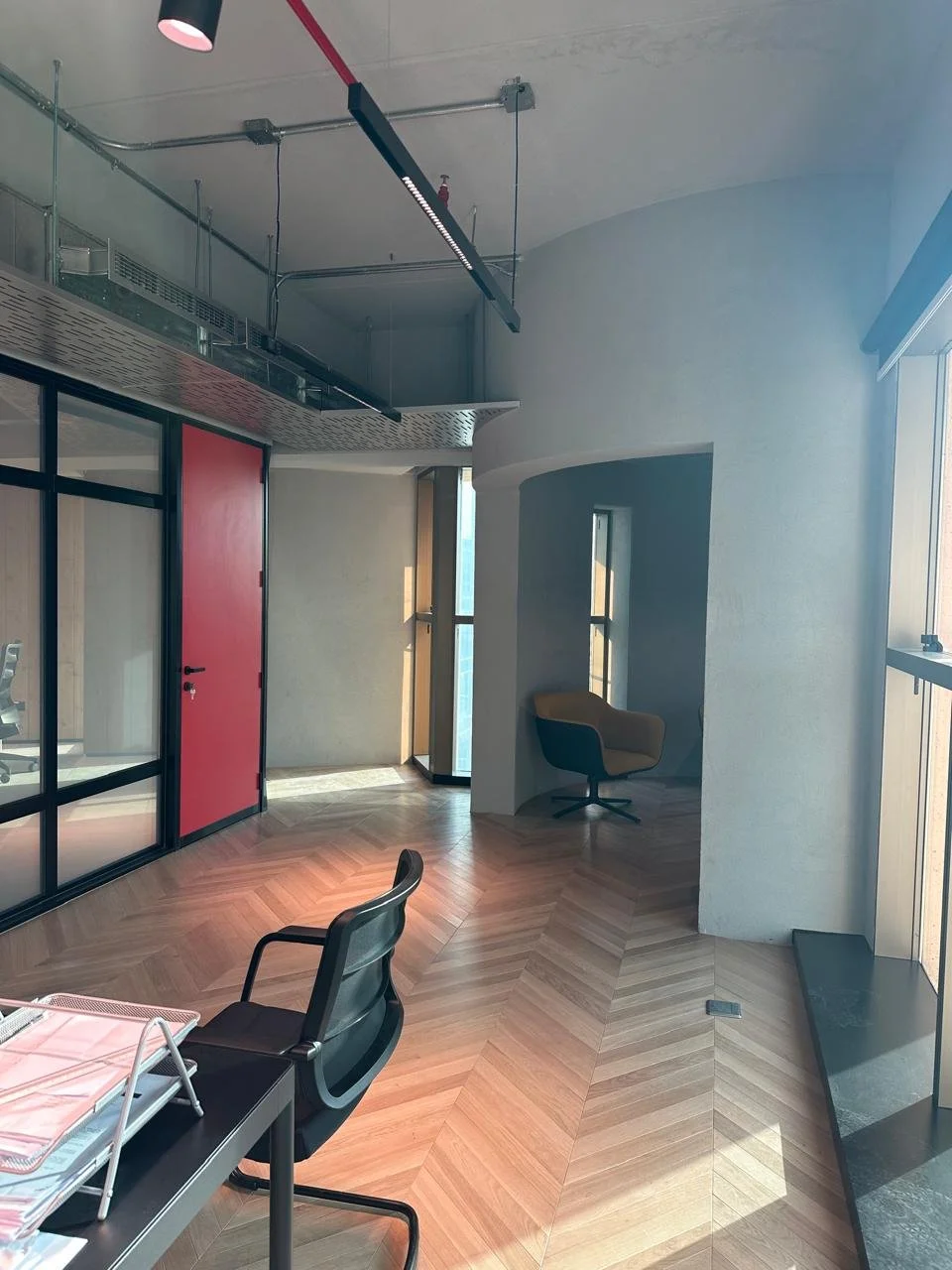 Modern office space with wooden chevron-patterned floor, black chairs, a red door, glass walls, and sunlight coming through large windows.