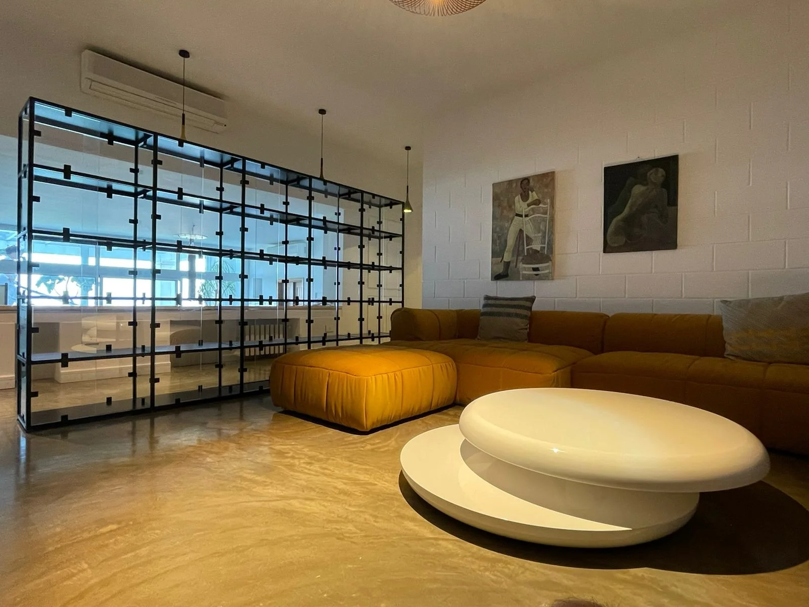 Modern living room with a large mustard-colored sectional sofa, a round white coffee table, and art on the white brick wall. A black metal room divider with glass panels separates the space from a view of a bright room with large windows and outdoor 