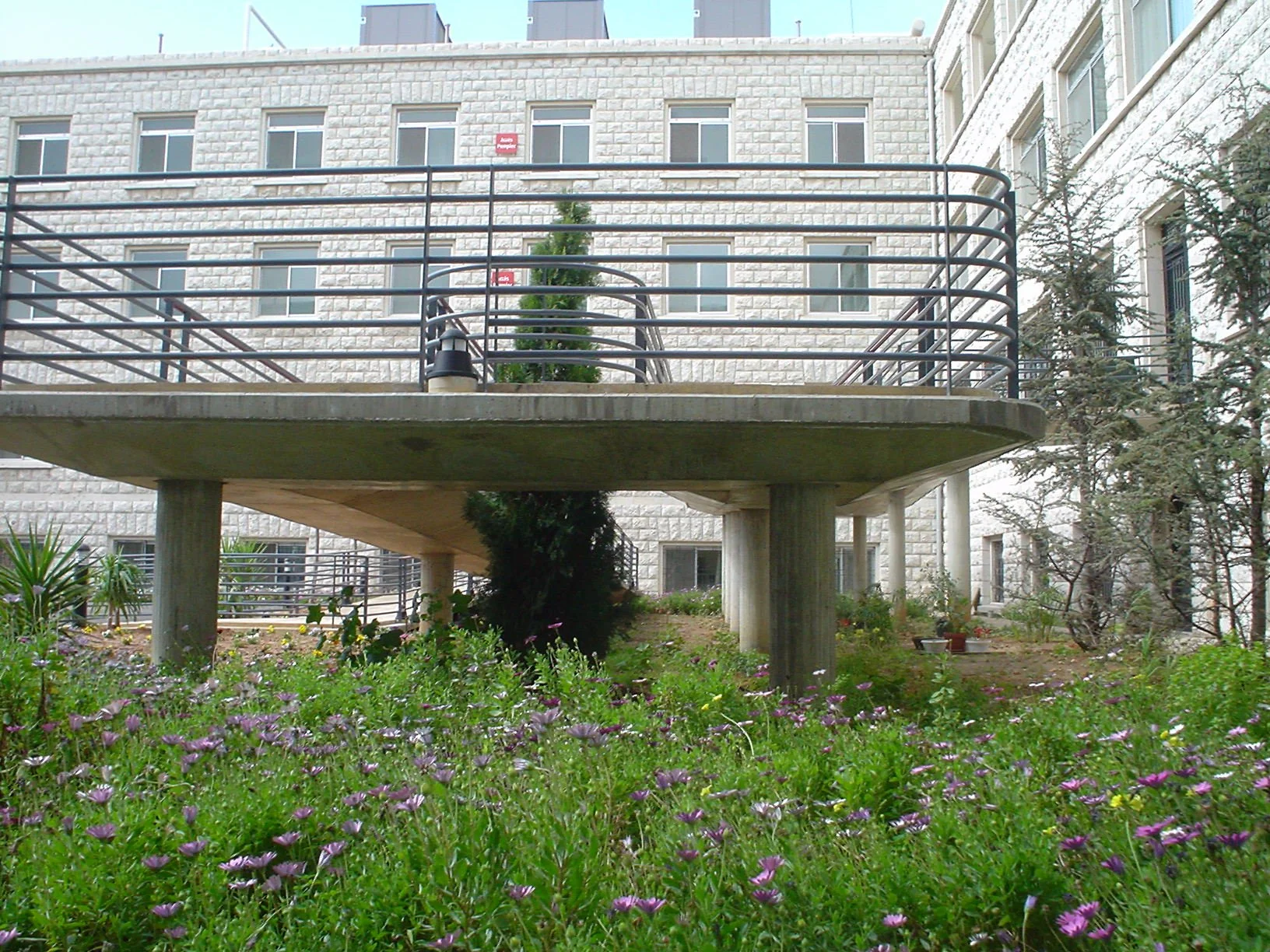 A modern residential building with a brick exterior, with a raised concrete balcony supported by pillars, surrounded by landscaped gardens with flowers and trees.