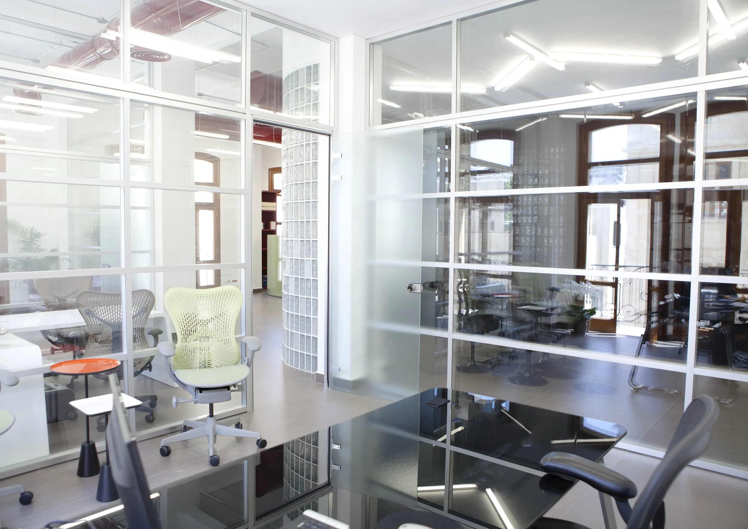 Modern office space with glass walls, white and gray office chairs, a black desk, and natural light coming through large windows.