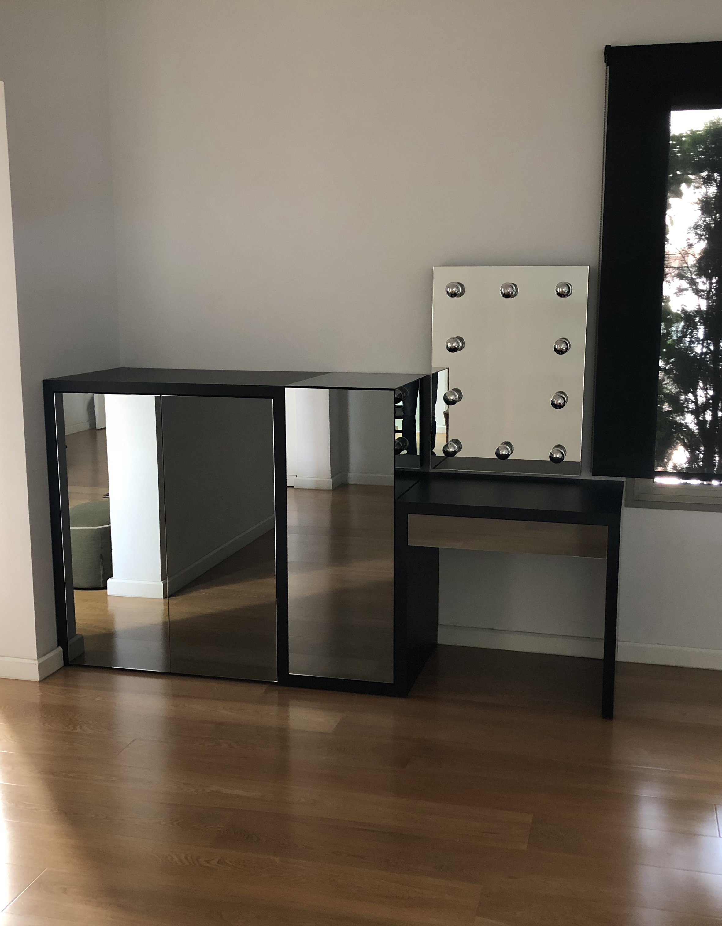 A modern, minimalist room with a black and white vanity mirror with lights, a black cabinet, and a window with a black curtain showing outdoor trees and sky.
