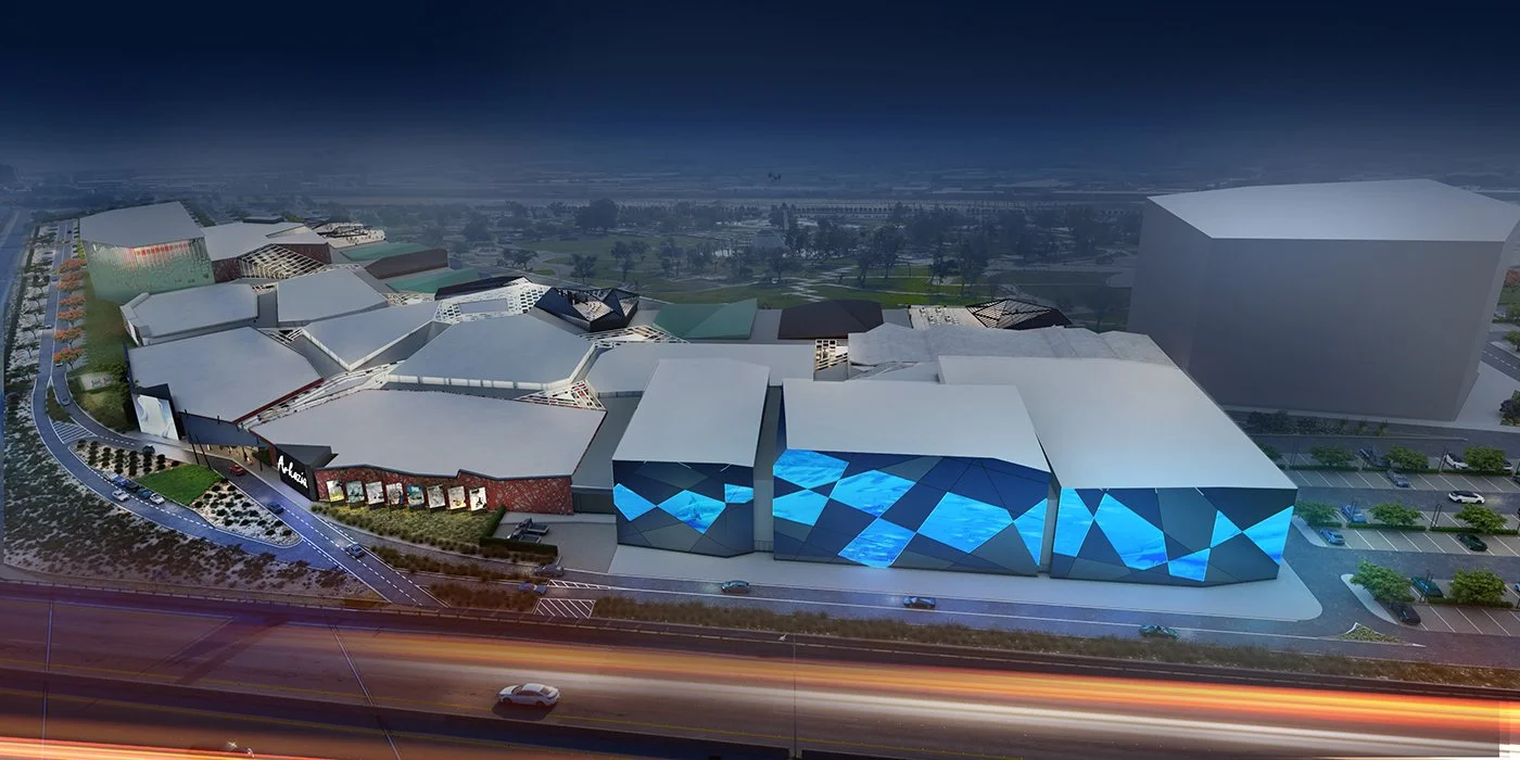 A digital rendering of a large modern shopping mall with unique geometrical roof designs and blue illuminated facades, situated near a highway with parking lots and surrounding greenery.