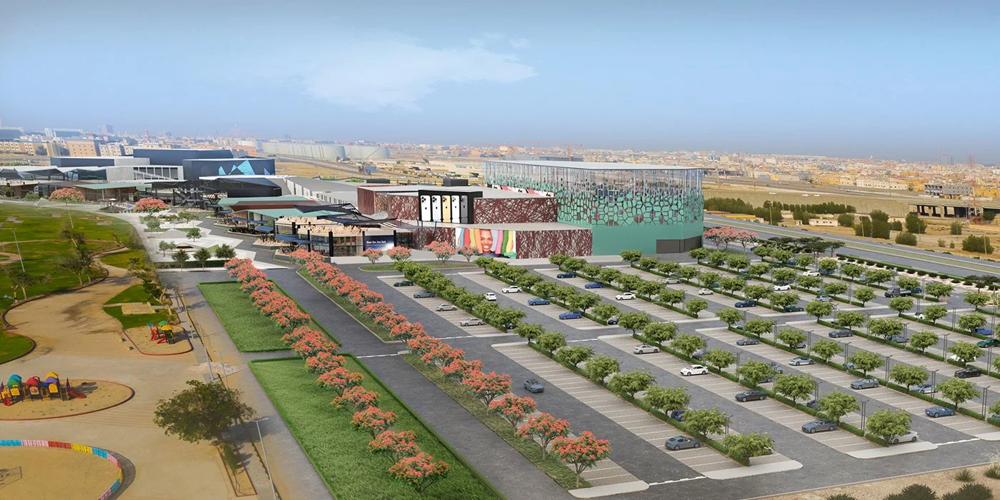 A large modern shopping mall with extensive parking lot, surrounded by landscaped green areas and colorful play areas, set in an urban environment with a distant cityscape in the background.