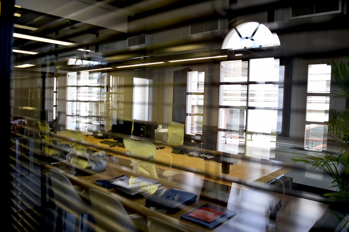 A modern office conference room with a long wooden table, yellow mesh chairs, multiple computer monitors, and large windows letting in natural light, viewed through window blinds.