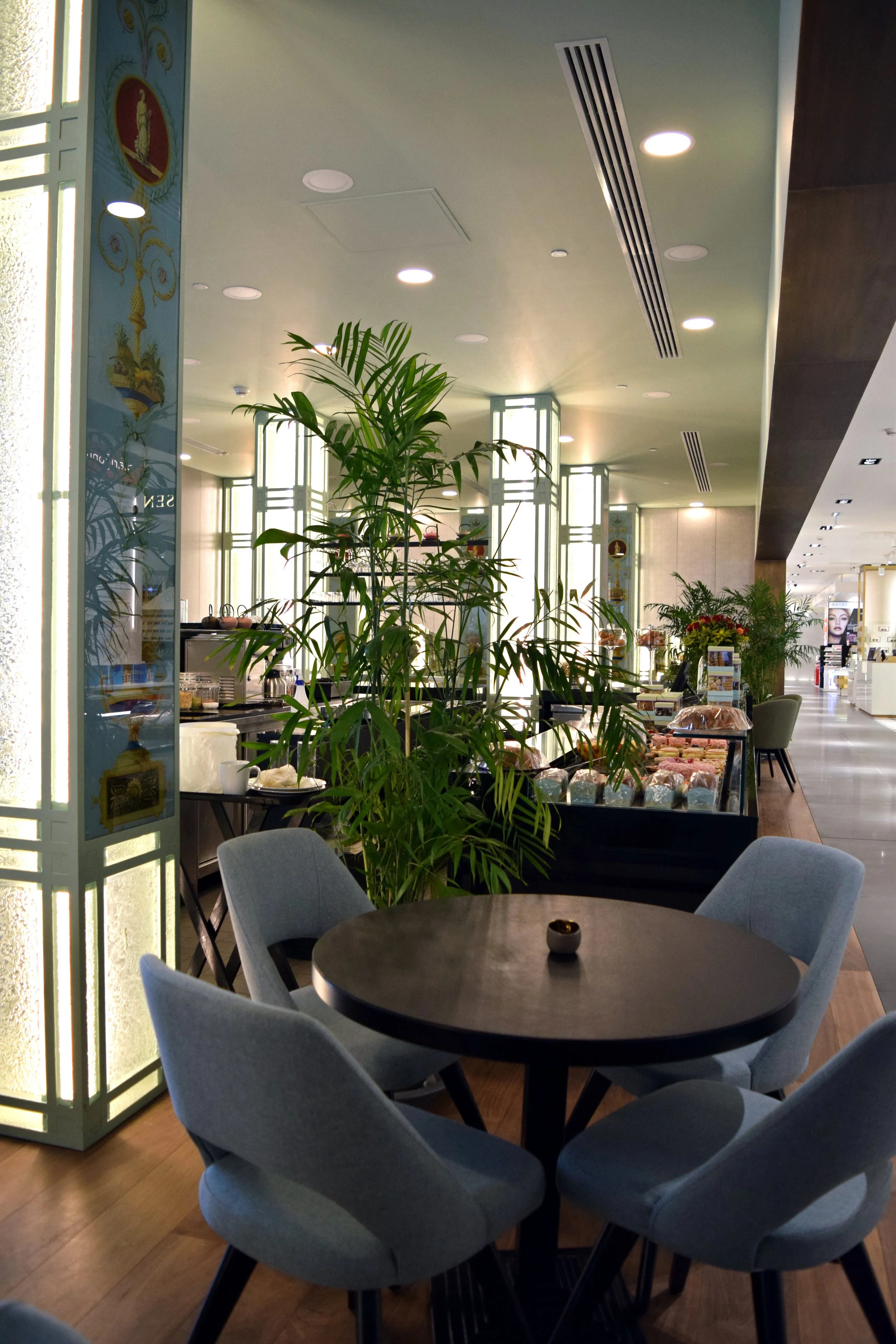 Indoor space with a round table surrounded by light grey chairs, green plants, and a display of baked goods, with soft lighting and modern decor.