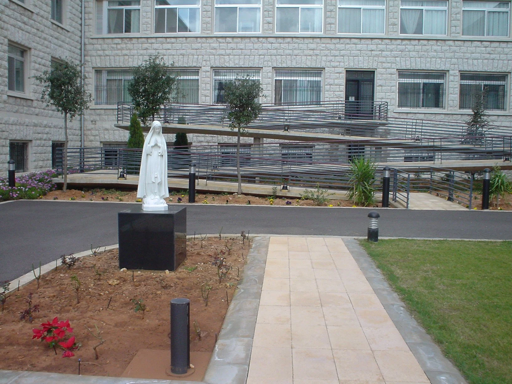 A courtyard with a paved pathway, a white statue of the Virgin Mary, small trees, and flower beds in front of a multi-story brick apartment building with large windows and a metal ramp.