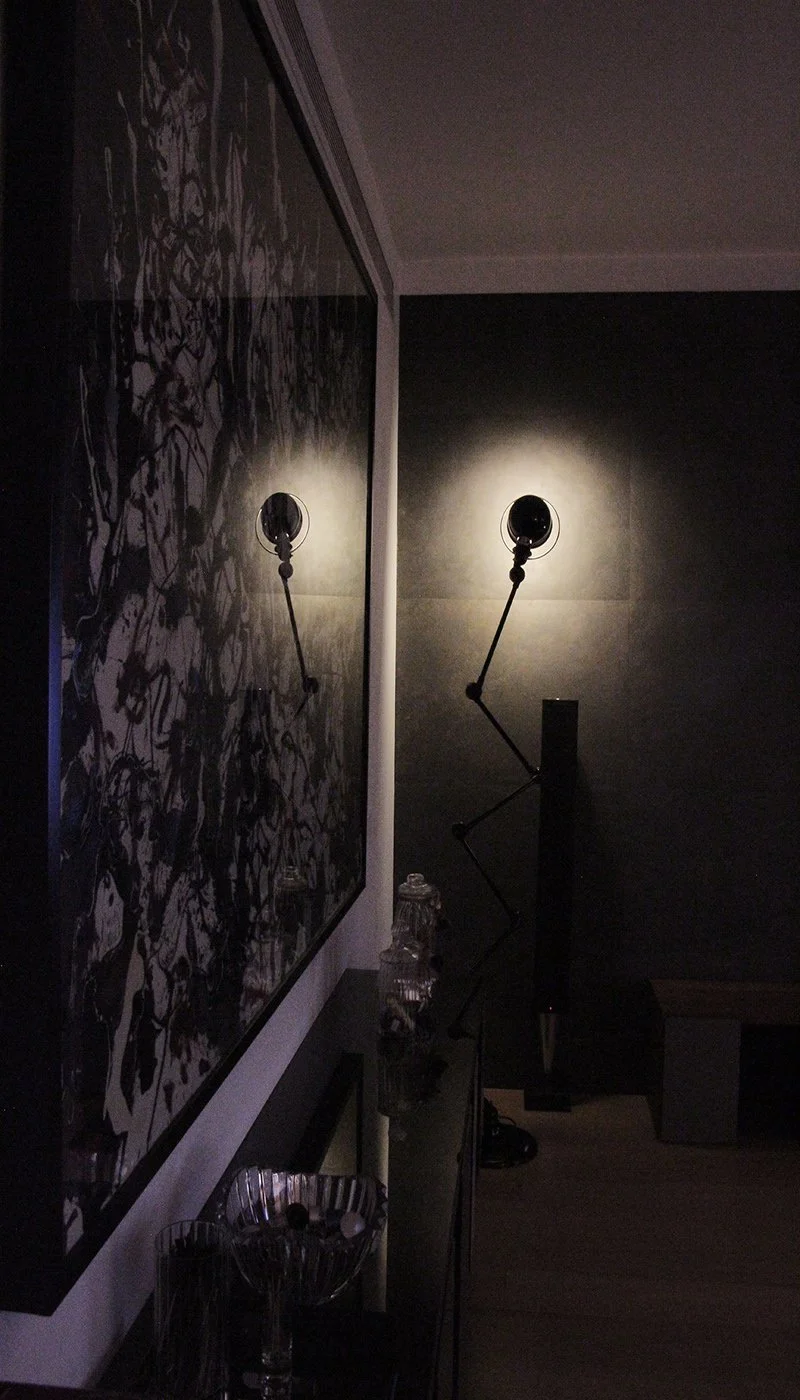 A dimly lit room with a modern wall-mounted lamp casting light on the dark wall, and a large wall art piece with a black and white abstract pattern. There are decorative items on a table below the art, including jars and glasses.