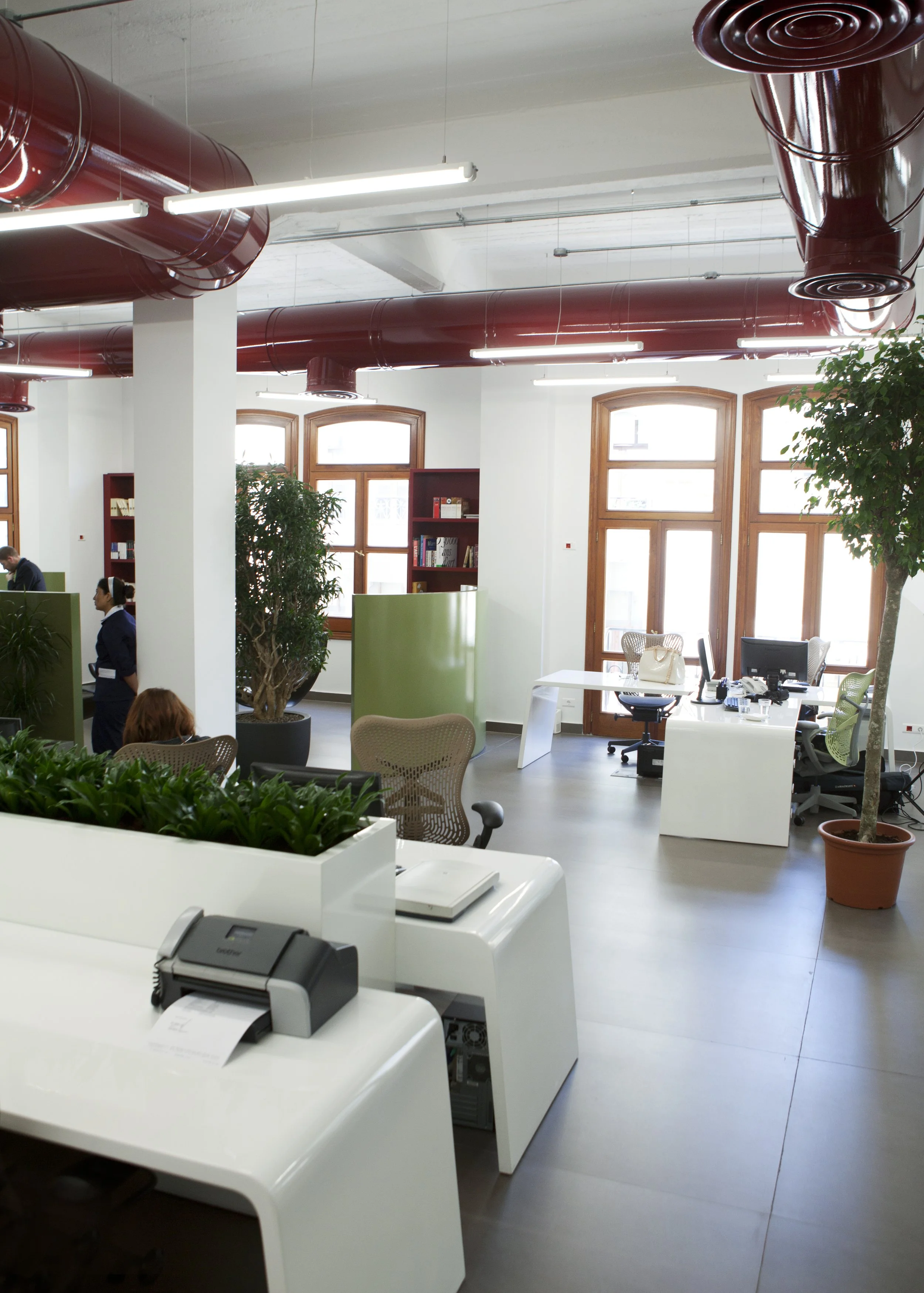 Modern office space with desks, office chairs, large windows, indoor plants, bookshelves, and exposed red ductwork on the ceiling.