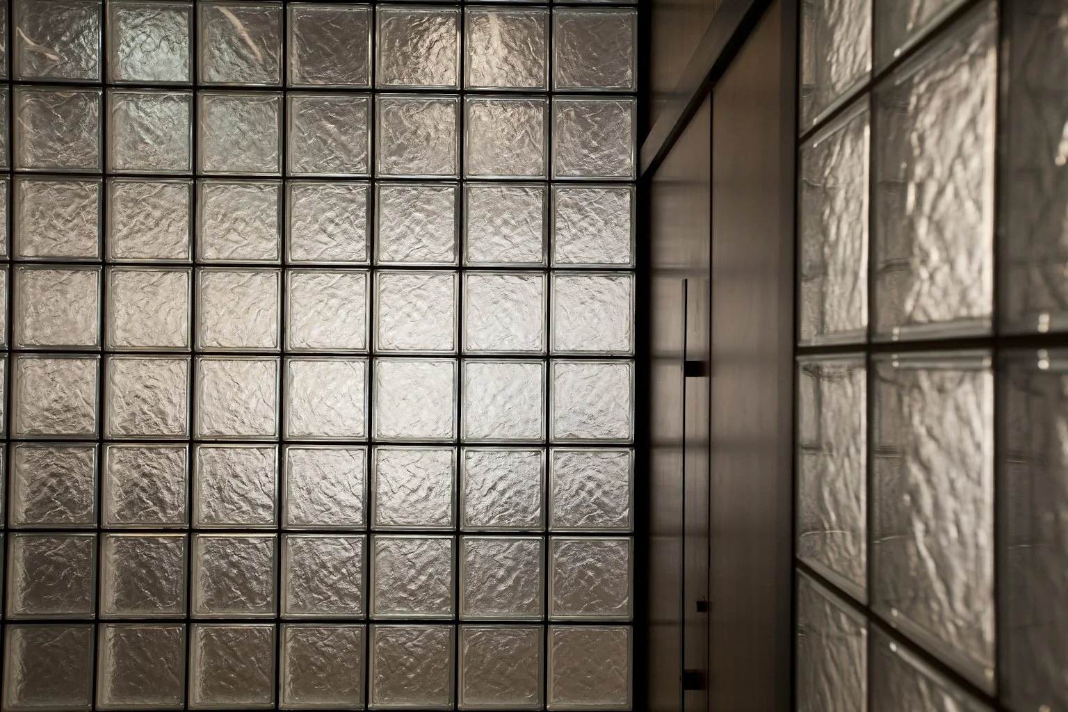 A wall made of glass blocks with a textured surface, separated by dark metal frames, with a wooden panel on the right side.
