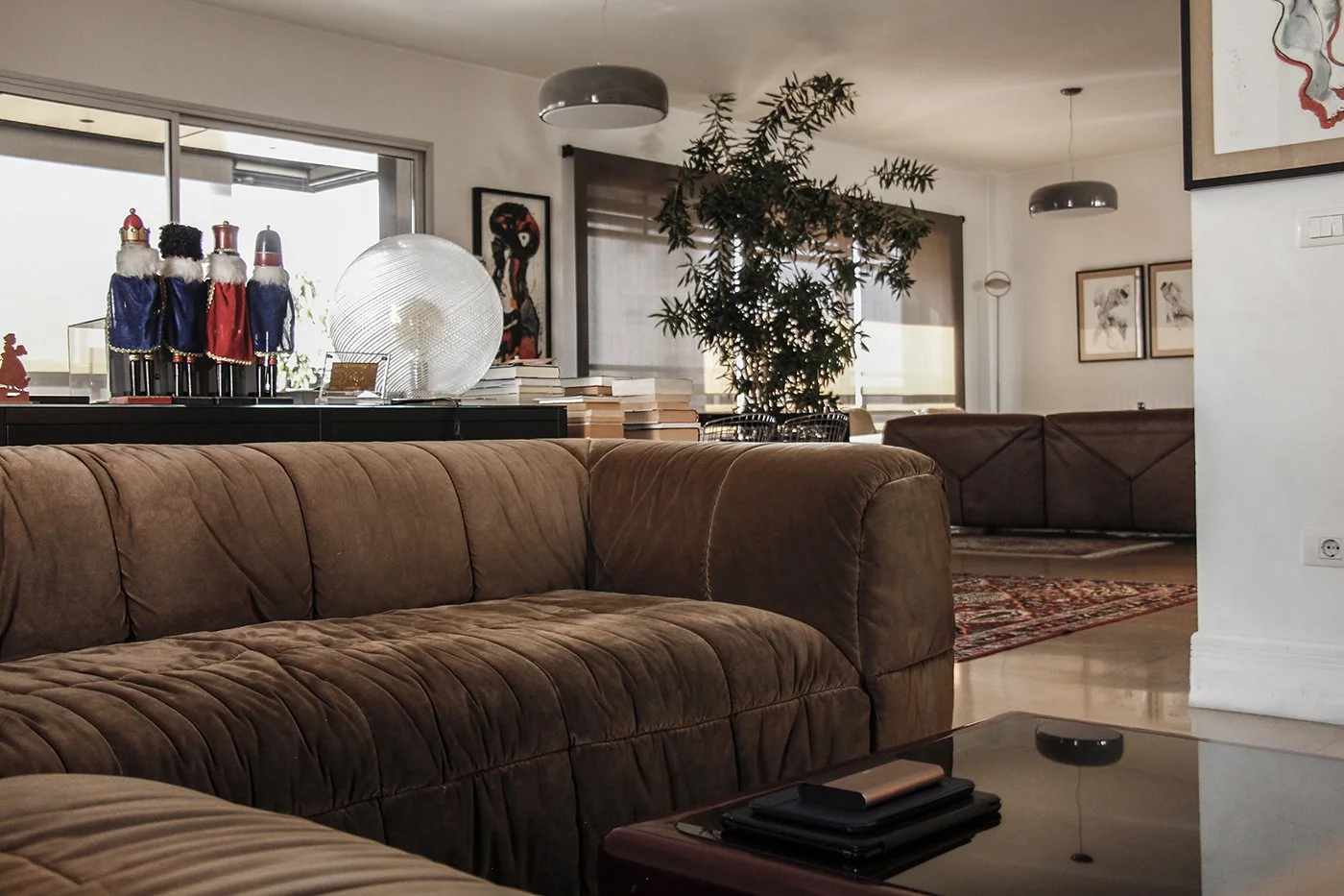 Living room with a brown velvet sofa, a coffee table with remote controls, Christmas decorations on a black cabinet, and artwork on the walls. A green potted plant is in the background near window blinds.