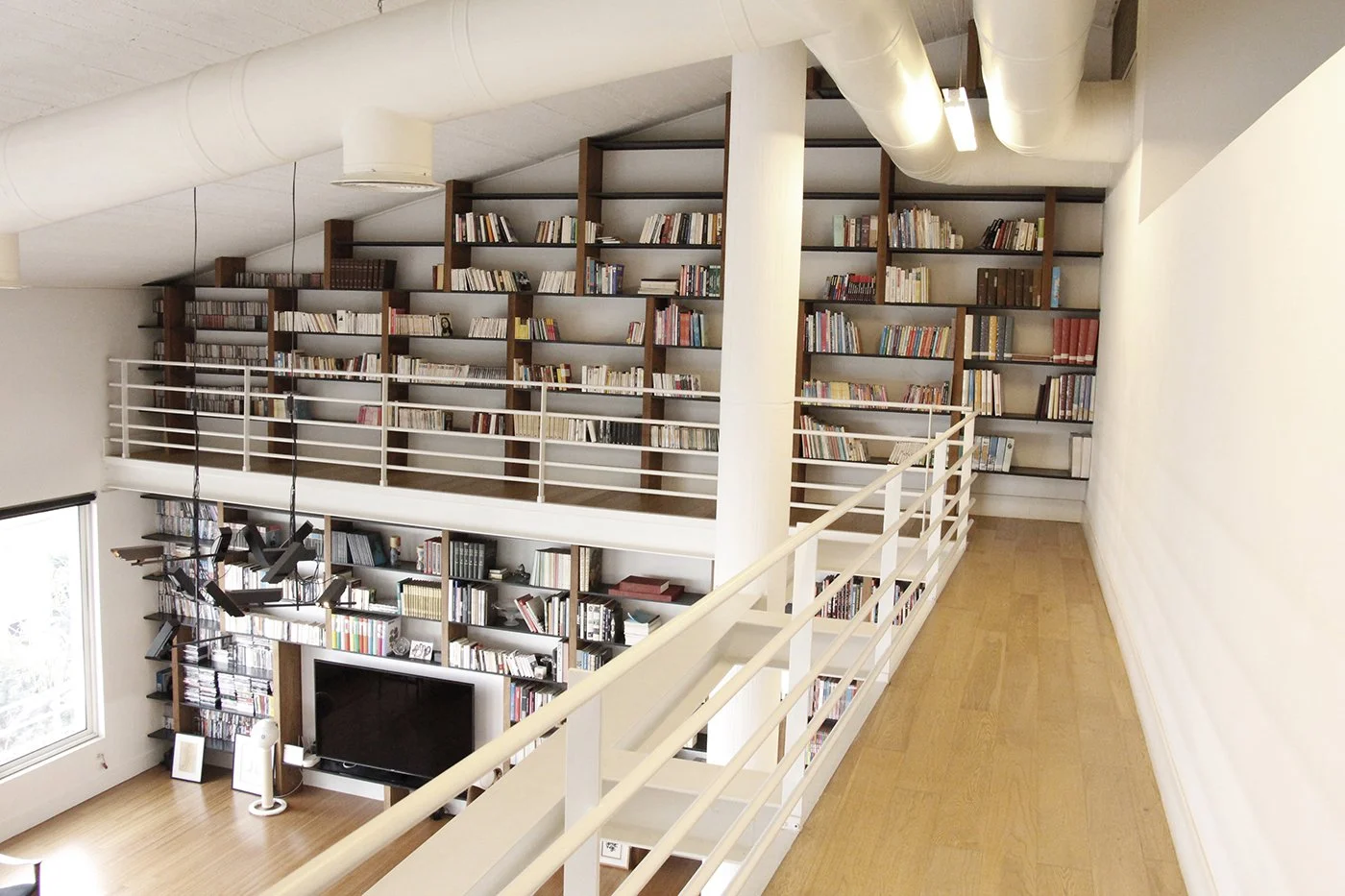 Interior view of a modern two-story house with a wooden floor, large bookshelf, and a flat-screen TV on the lower level, seen from an upstairs walkway.