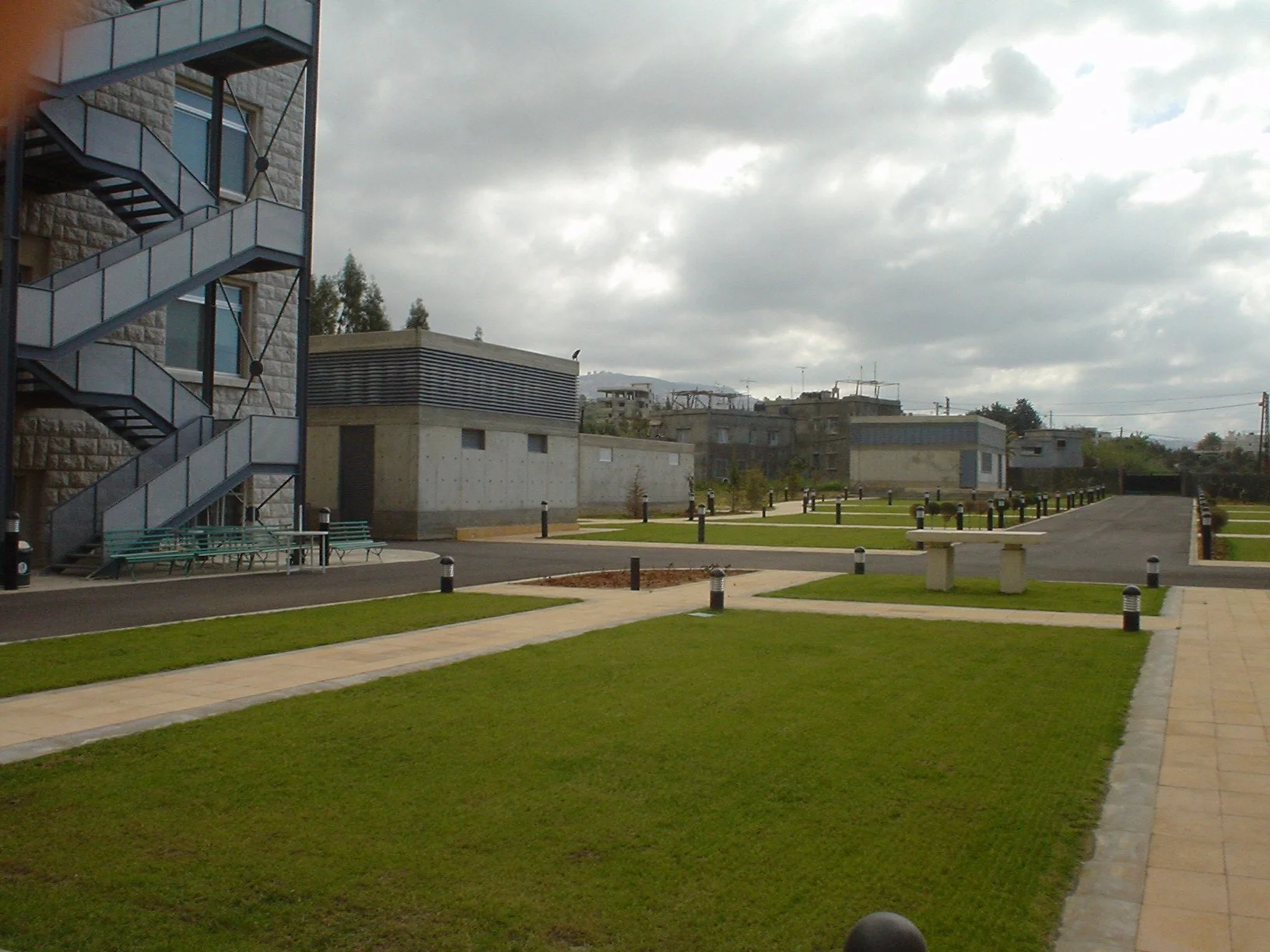 View of a modern residential area with green lawns, paved walkways, benches, and buildings with external staircases and flat rooftops under a cloudy sky.
