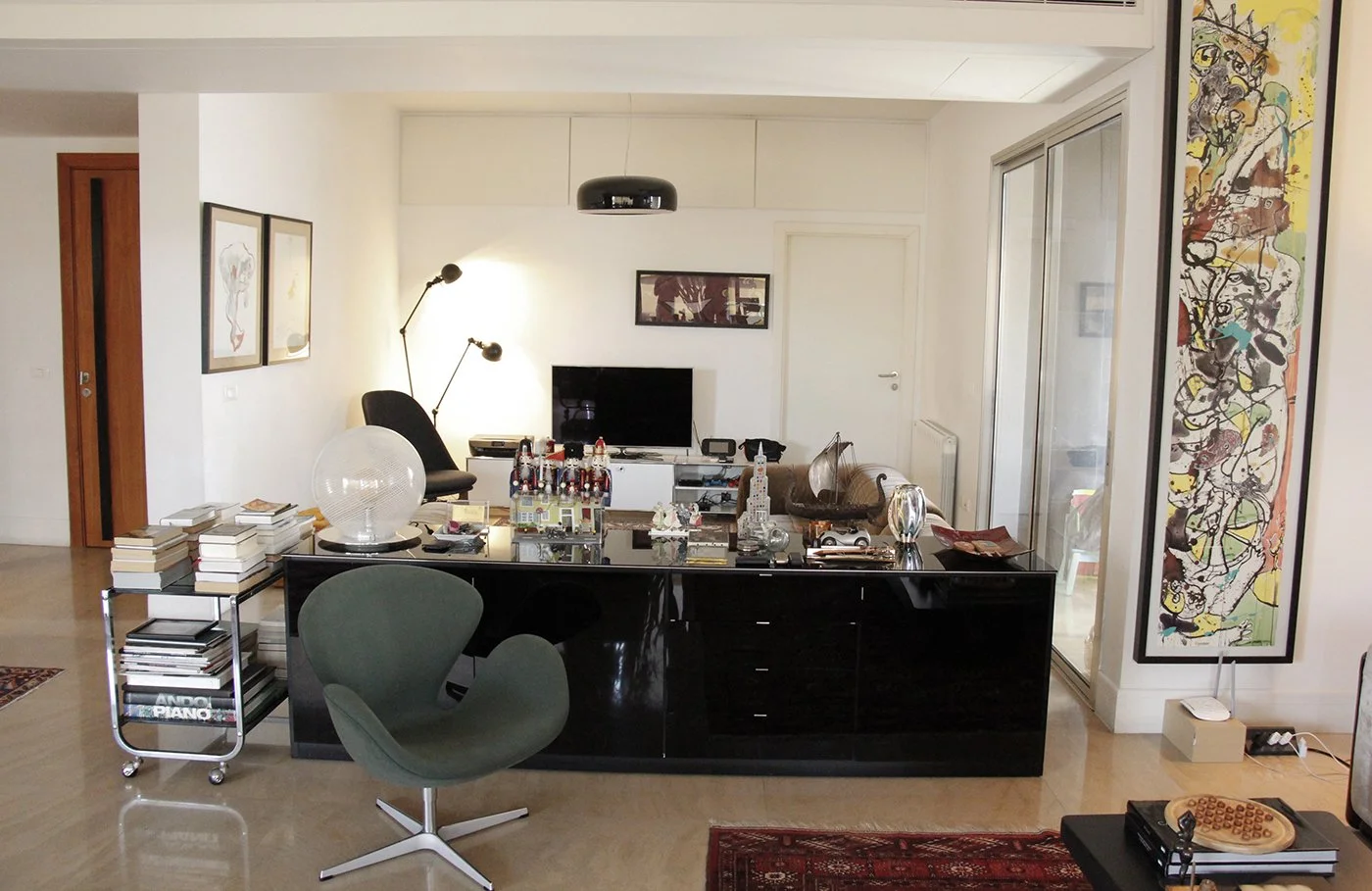 Living room with a black sideboard topped with books, decorations, and a large lamp. Behind is a TV, artwork on the white walls, and a glass sliding door. There are two chairs, one green and one black, and various decorative items and artwork through