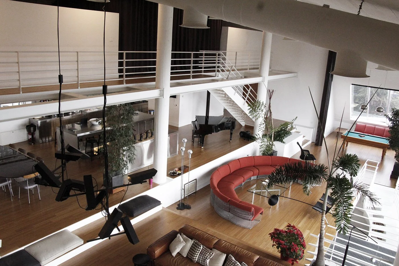 A spacious, modern loft-style living area with high ceilings, a curved reddish-orange sofa, black grand piano, and multiple potted plants. There's a staircase leading to an upper level, large windows letting in natural light, a pool table in the corn