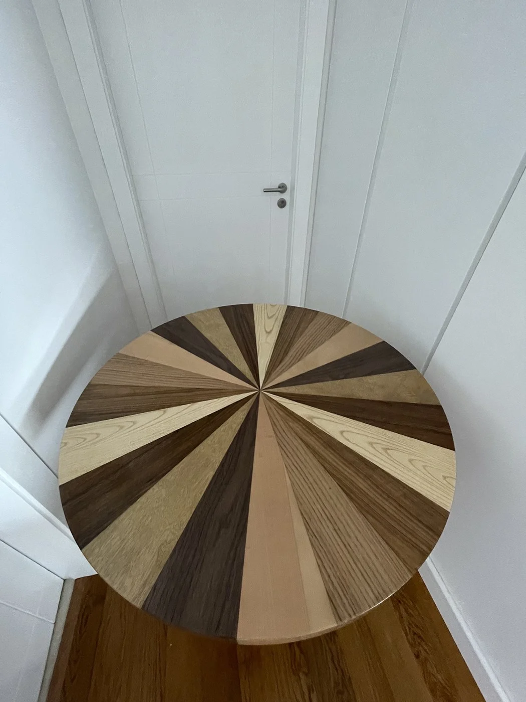 Round wooden table with a geometric pattern made from various shades of wood pieces in a room with white walls and a closed door.