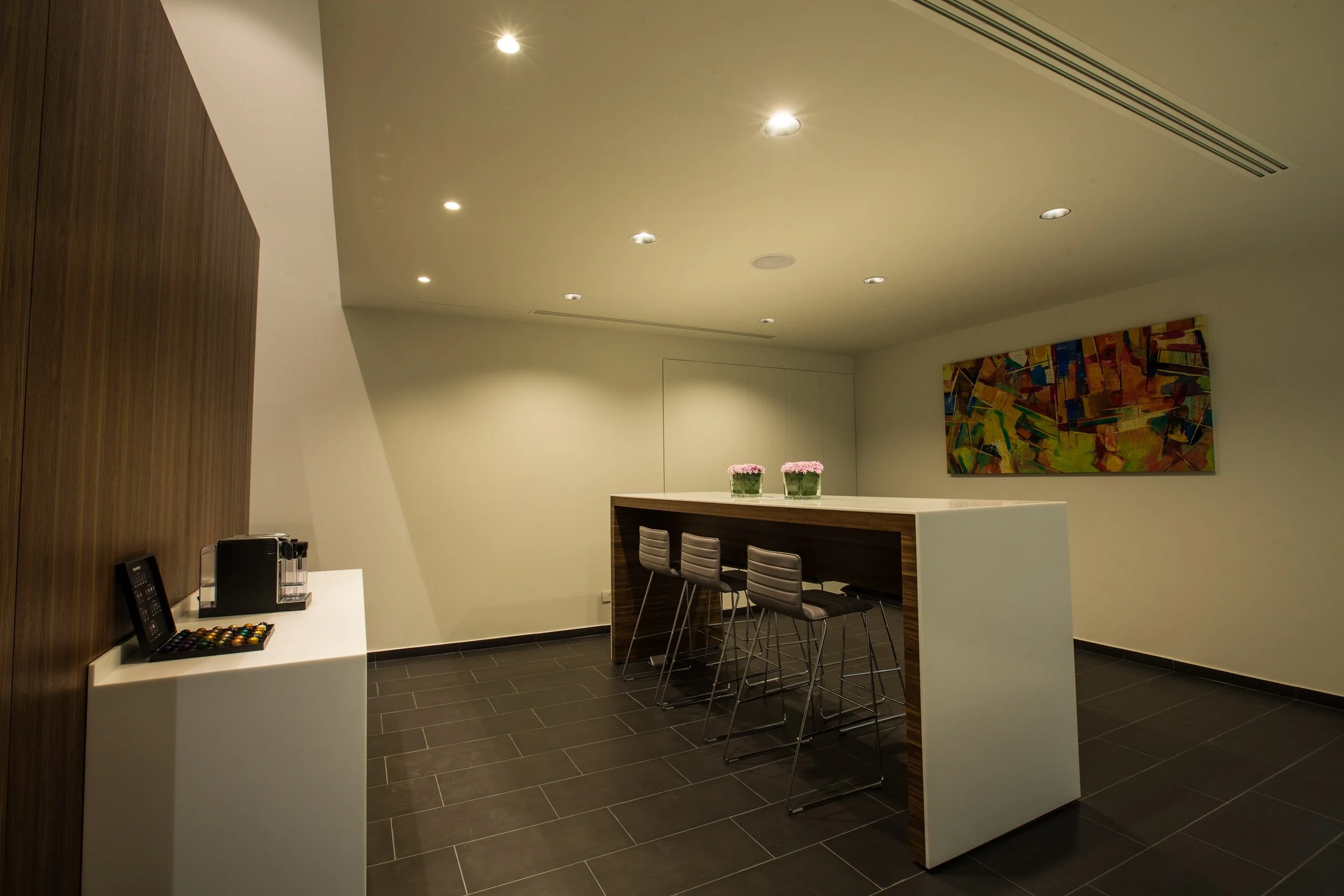 A modern hotel or office lounge with a high ceiling, recessed lighting, dark tiled floor, and a white and wood bar-style counter with four metal stools. Artwork with colorful geometric shapes hangs on the wall, and small pink flowers in glass contain