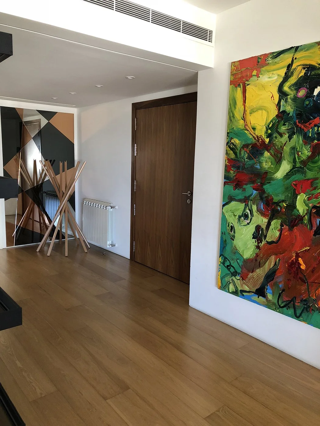 A modern interior space with wooden floors, white walls, a large colorful abstract painting on the right, a closed wooden door in the center, a radiator beneath a corner, and a decorative wooden coat rack with angled sticks on the left.