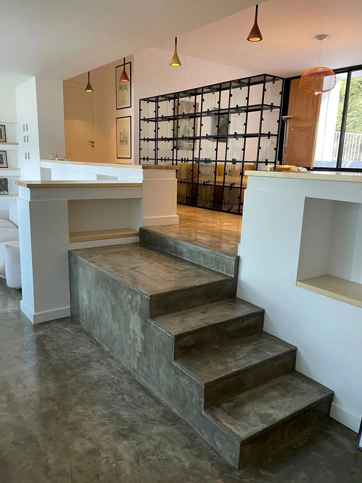 Modern interior with concrete stairs leading to a raised living area separated by a black metal grid partition. The space features white walls, wooden accents, pendant lighting, a window with a view of greenery, and artwork on the walls.