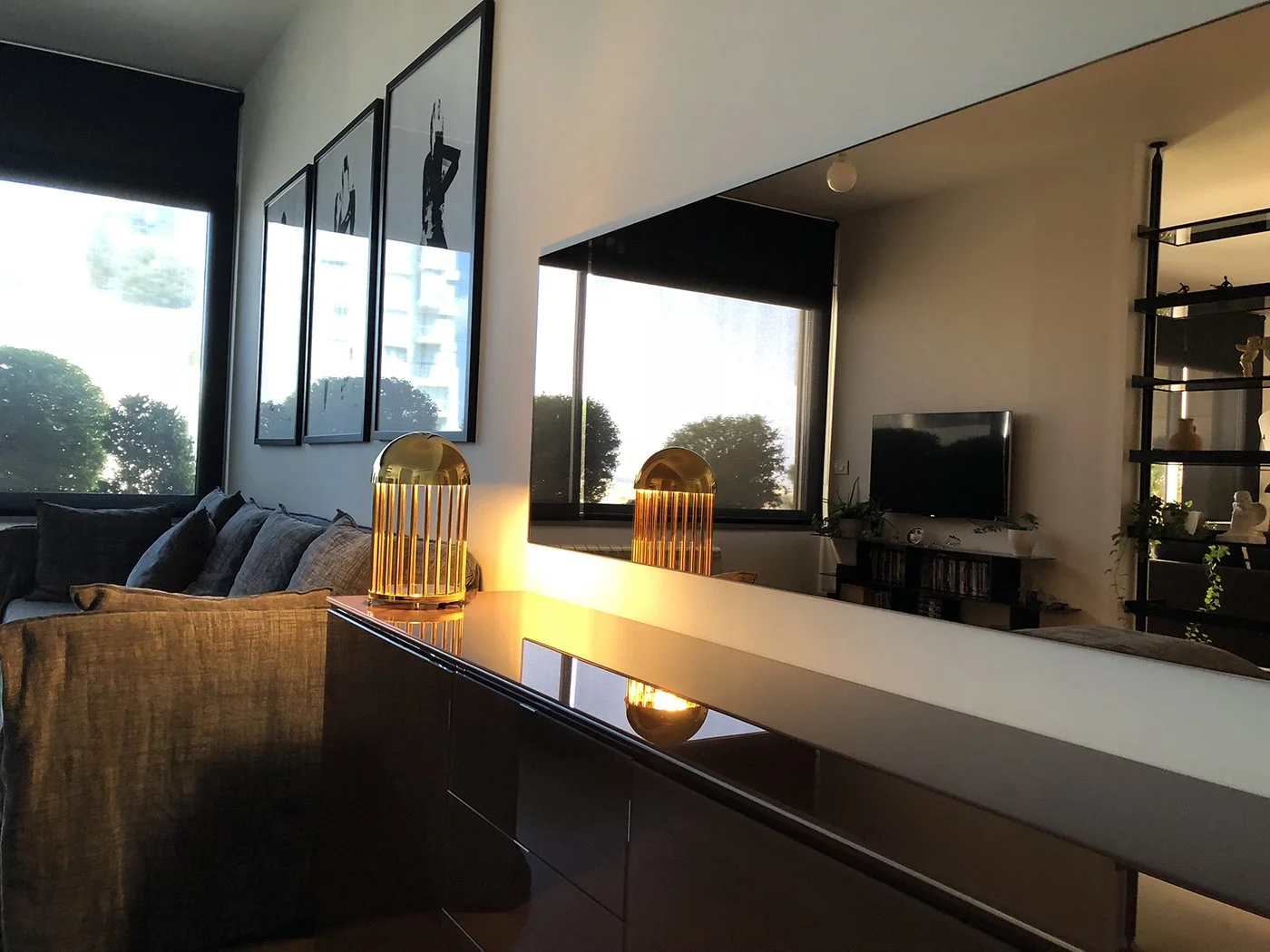 Modern living room with large windows, a black TV on a stand, decorative plants, black shelves with ornaments, and a dark wood dining table with two gold lamps.
