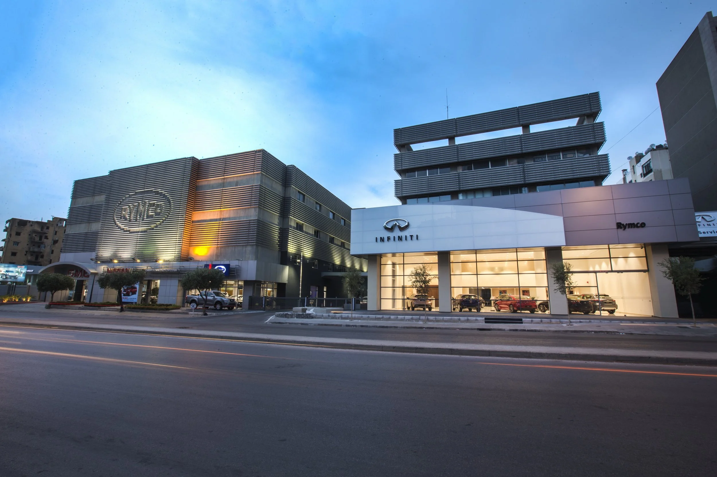 A city street scene at dusk showing a modern car dealership building with the INFINITI logo, adjacent to other commercial buildings, with cars parked in front and some trees lining the sidewalk; the sky is partly cloudy with a hint of sunset light.