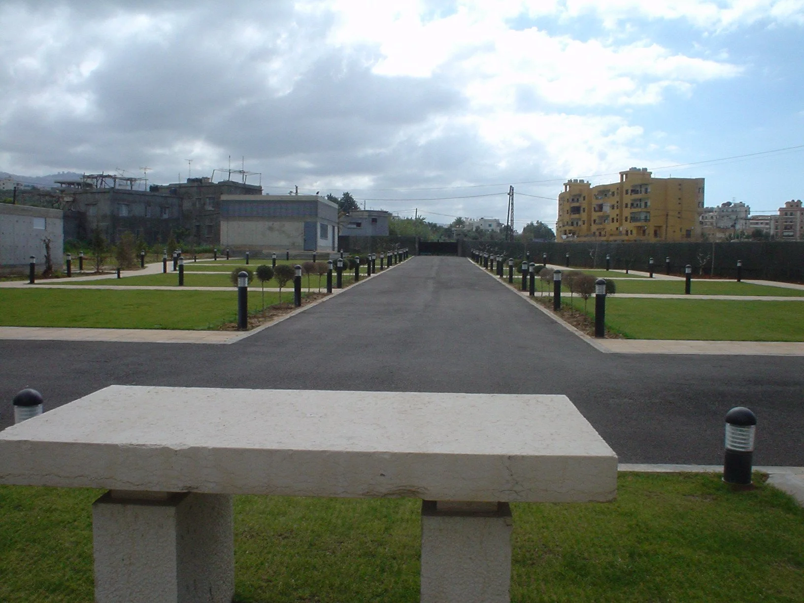 A park with a concrete picnic bench in foreground, green grass, black pathway, lampposts, trees, and multistory buildings in the background under cloudy sky.