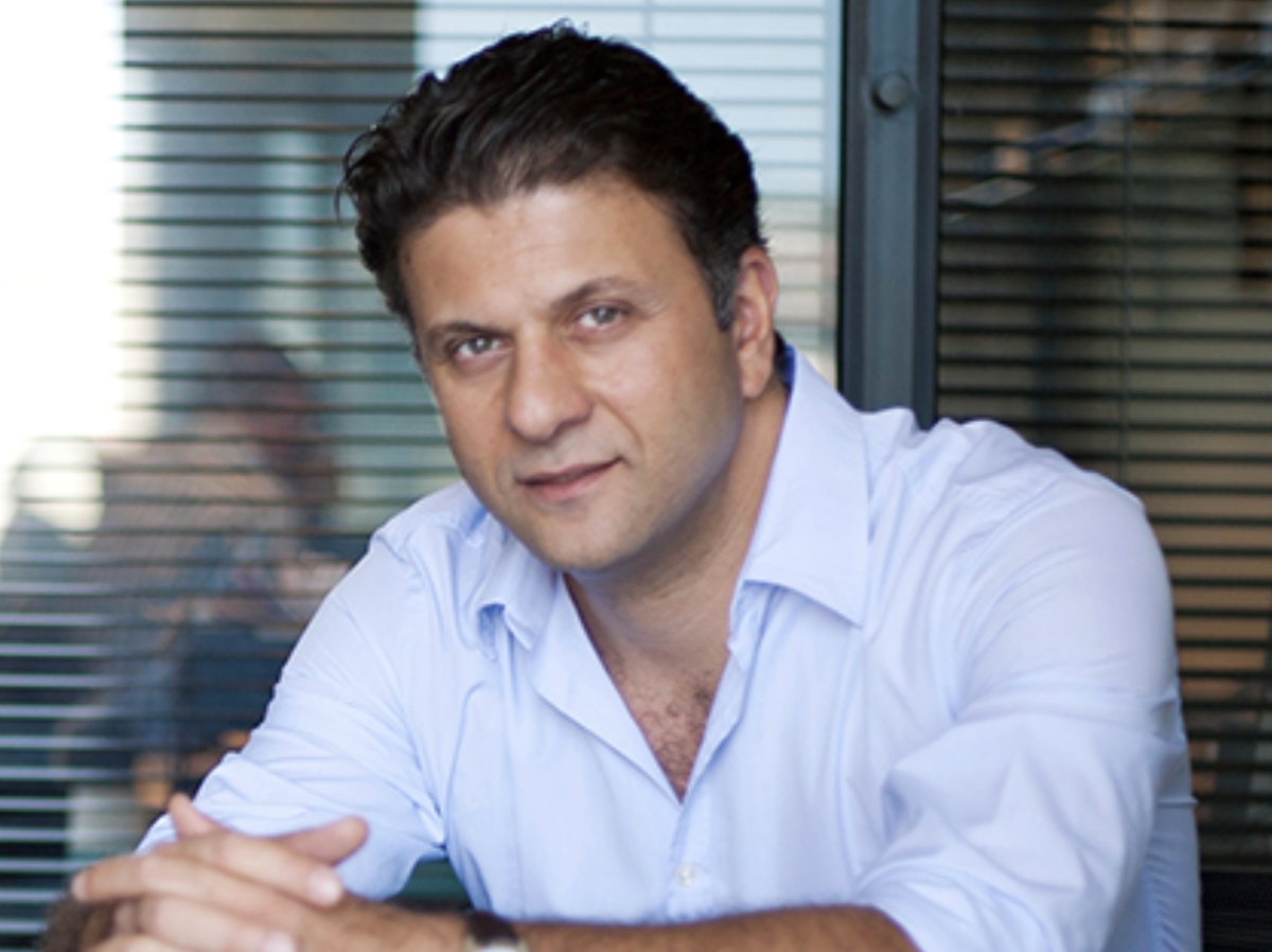 A man with dark hair and a light blue dress shirt sitting in front of glass walls with blinds, looking at the camera.