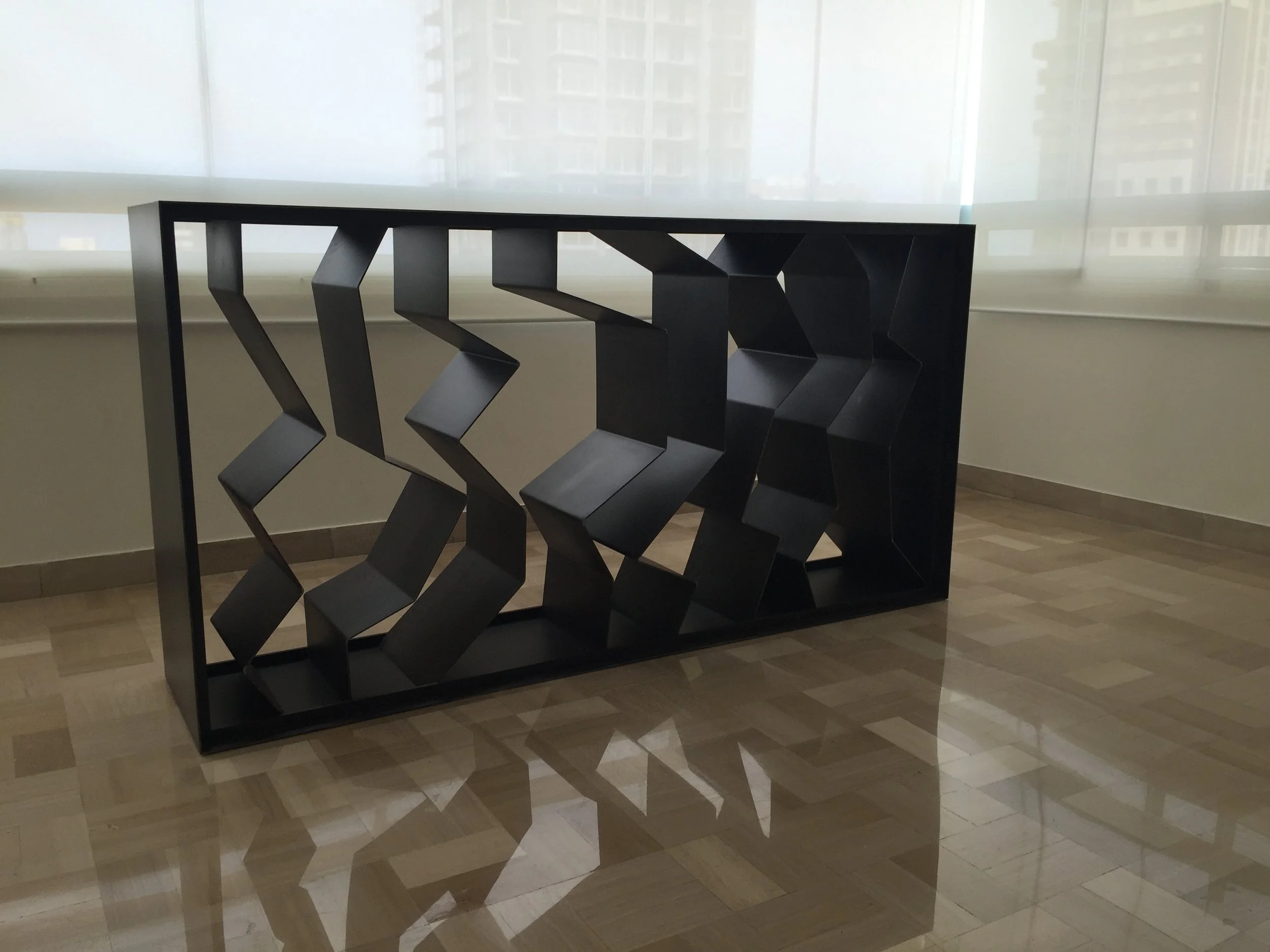 A black geometric sculpture with multiple angular, interconnected sections, placed on the floor near a large window with sheer blinds.