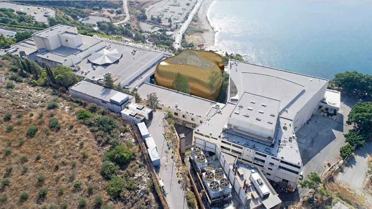 Aerial view of a modern building with a gold-colored, organic-shaped structure on the rooftop, near a coastline with water and a beach.