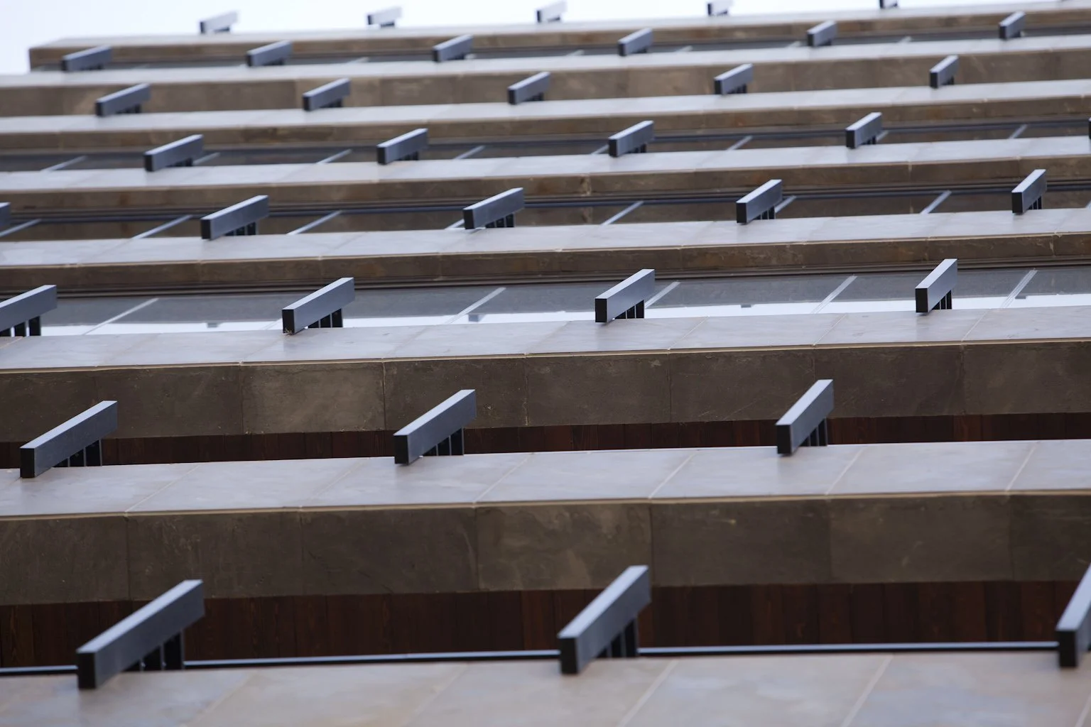Multiple rows of building balconies with dark metal railings and brown stone or concrete floors, viewed from below.