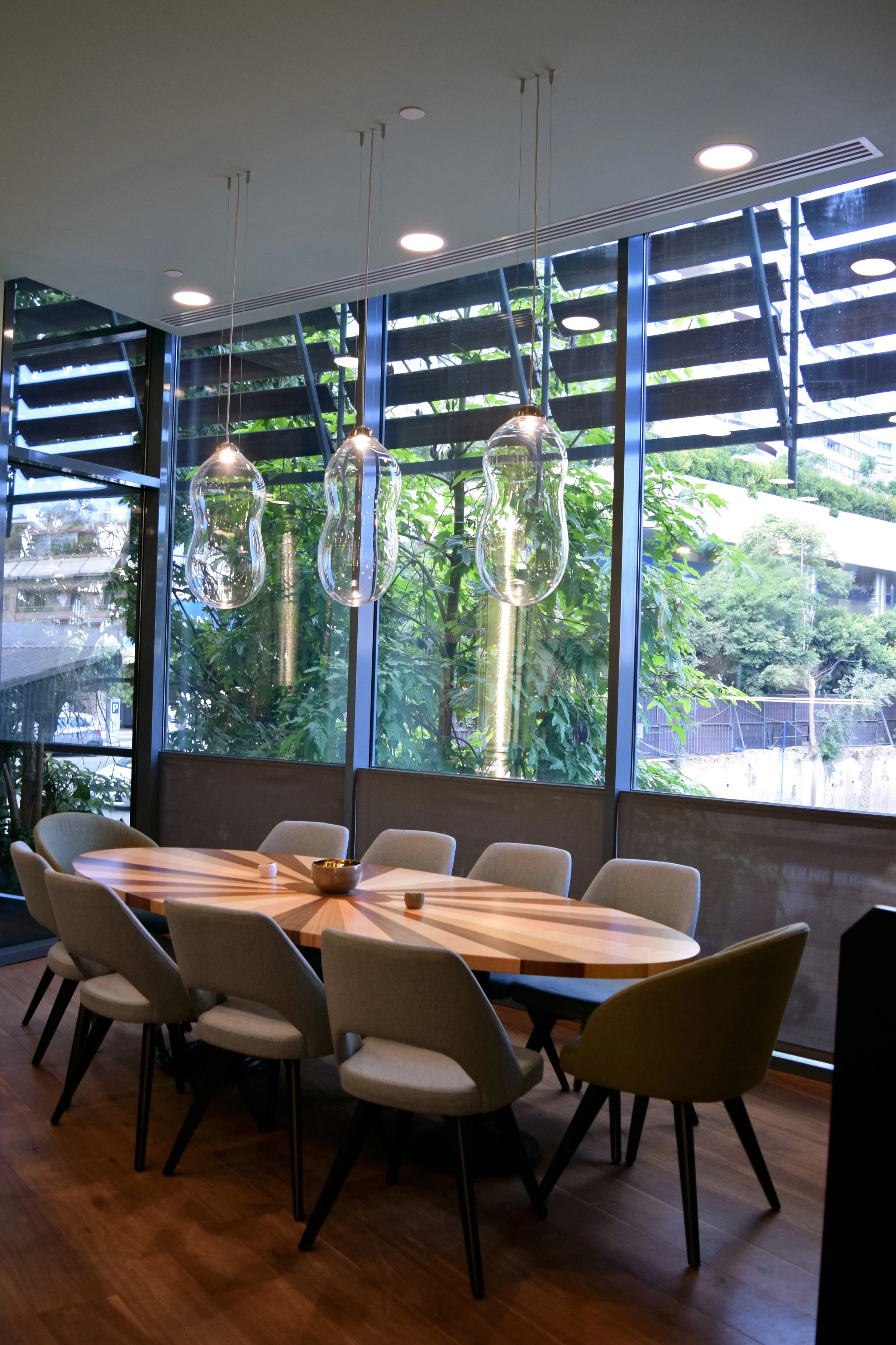 Modern dining area with a wooden table, beige chairs, large windows with greenery outside, hanging glass pendant lights, and a wooden floor.