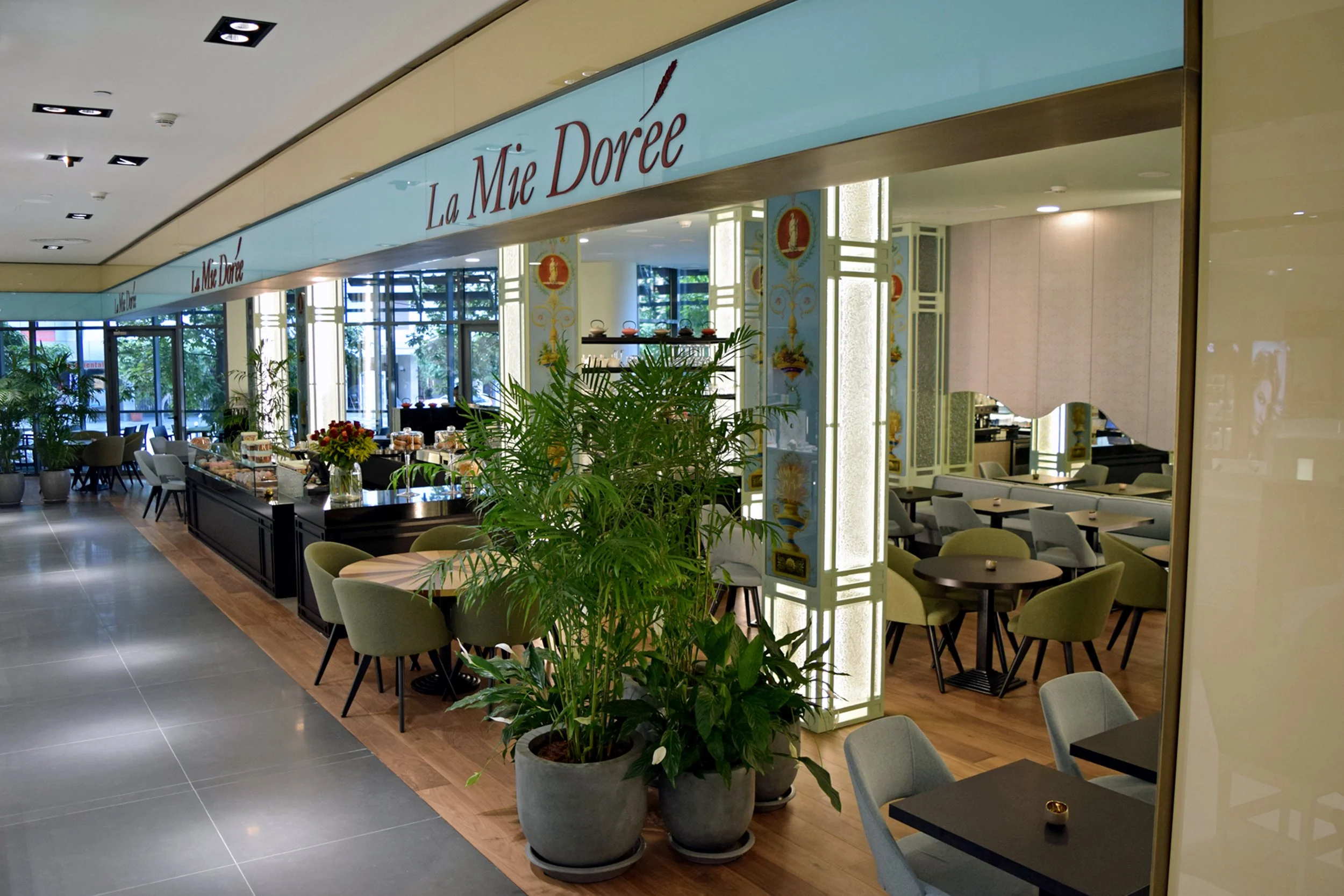 Interior of a restaurant or café named La Mie Dorée, with green chairs, dark tables, potted plants, and large windows, decorated with elegant patterns and a lit display area.