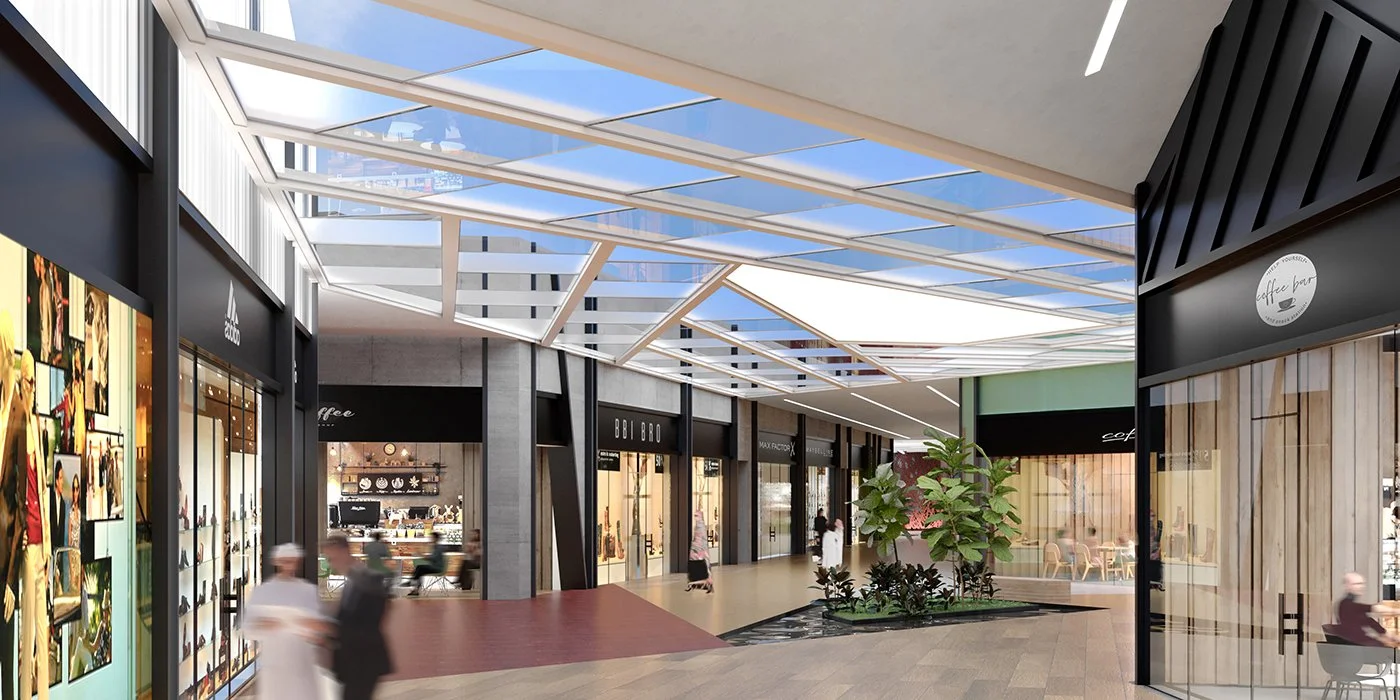 Interior view of a modern shopping mall with storefronts, large glass ceiling, and a seating area with plants.
